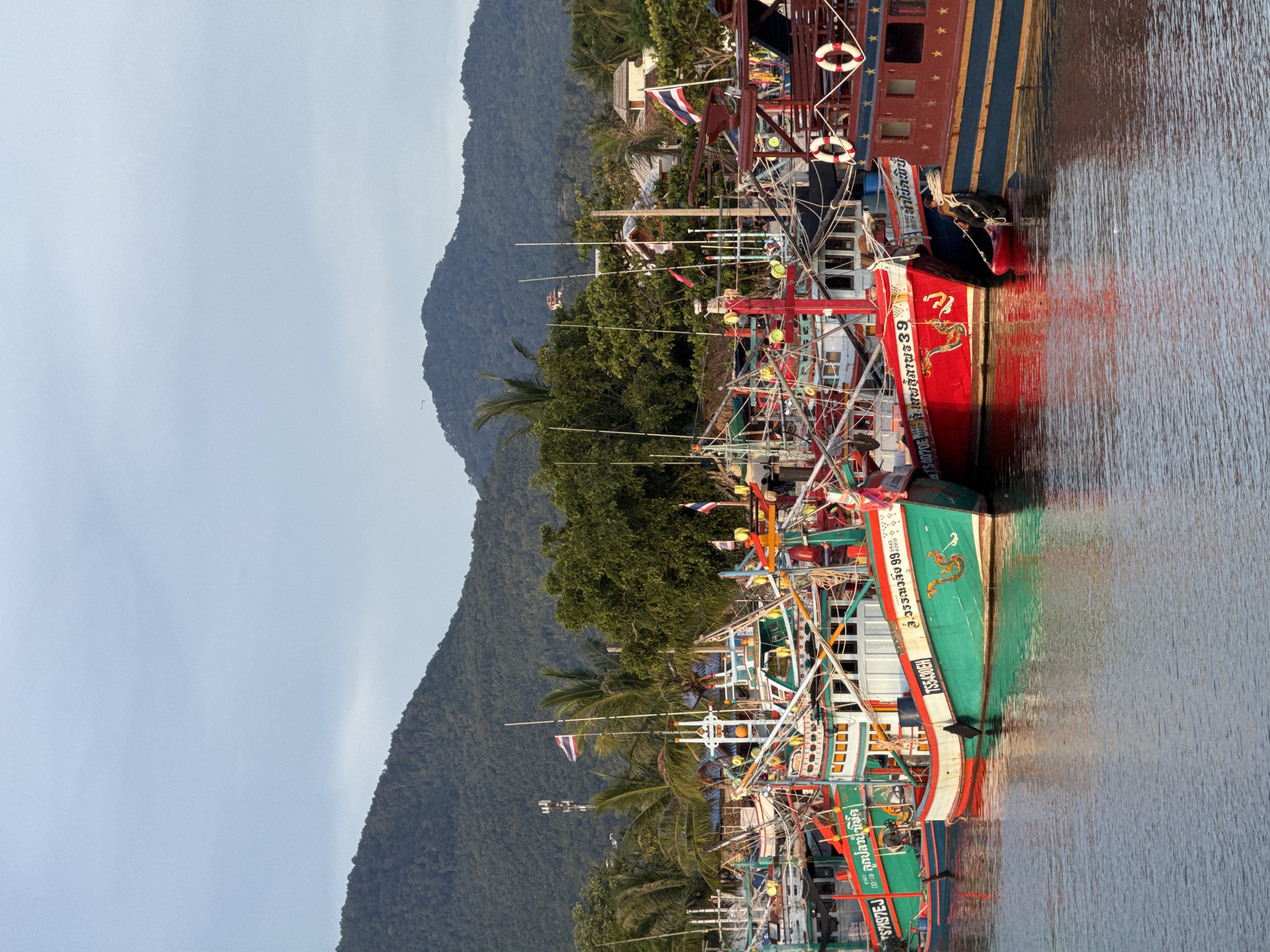 The image depicts a harbor in Koh Phangan, Thailand, with several fishing boats moored together. The boats are of various sizes and colors, with the majority being green and red. Many of them are decorated with intricate designs and lettering in Thai script. A few Thai flags are visible, one flying from a tall mast. In the background, lush green mountains rise, covered in dense vegetation. The sky is a pale blue with some wispy clouds. The water in the harbor is calm, reflecting the boats and the surrounding scenery. A person dressed in dark clothing is visible near the right side of the image, tending to ropes connected to one of the boats. The overall scene suggests a working harbor in a tropical coastal region.