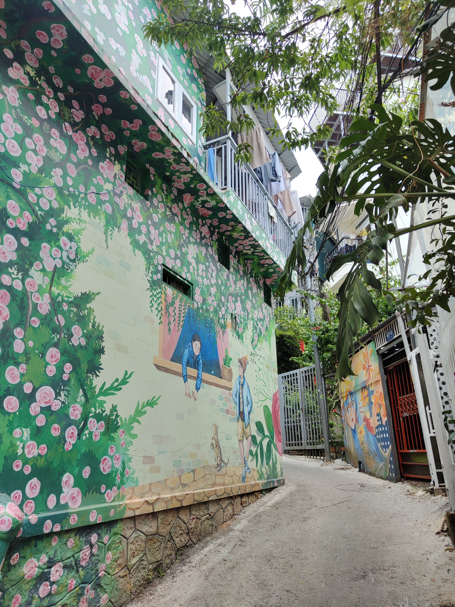 A narrow alleyway in Da Lat, Vietnam, is flanked by buildings adorned with vibrant murals. On the left, a multi-story building features a mural of pink flowers and figures, including a man leaning out of a painted window and another standing with a small animal. The building's exterior is decorated with a repeating flower pattern. A stone-like base runs along the bottom of the building. On the right, a smaller building displays a colorful mural of people and flowers. A metal gate and an orange door are visible further down the alley. Utility wires crisscross overhead, partially obscured by foliage. The alleyway itself is paved.