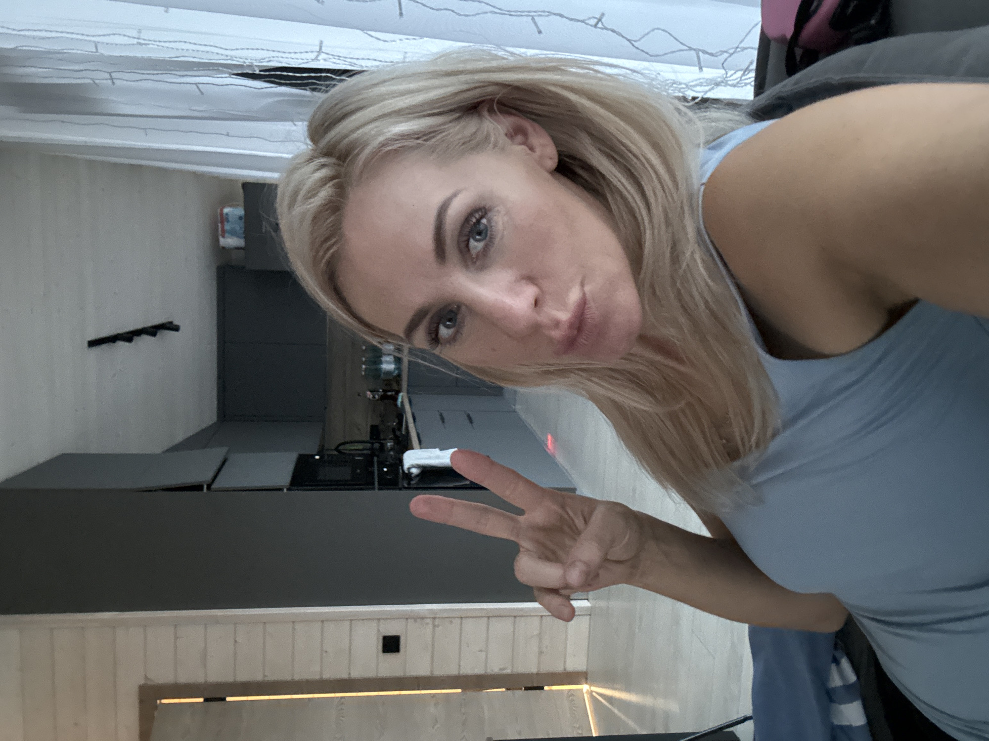 A woman with blonde hair is taking a selfie in a modern living room. She is wearing a light blue top and making a peace sign with her left hand. The background shows a kitchen with dark gray cabinets, a wooden countertop, and a black stove. There's also a window with sheer white curtains and string lights. The room has a minimalist aesthetic with wooden paneling on the walls and a light-colored floor. The overall impression is one of a comfortable, contemporary home.