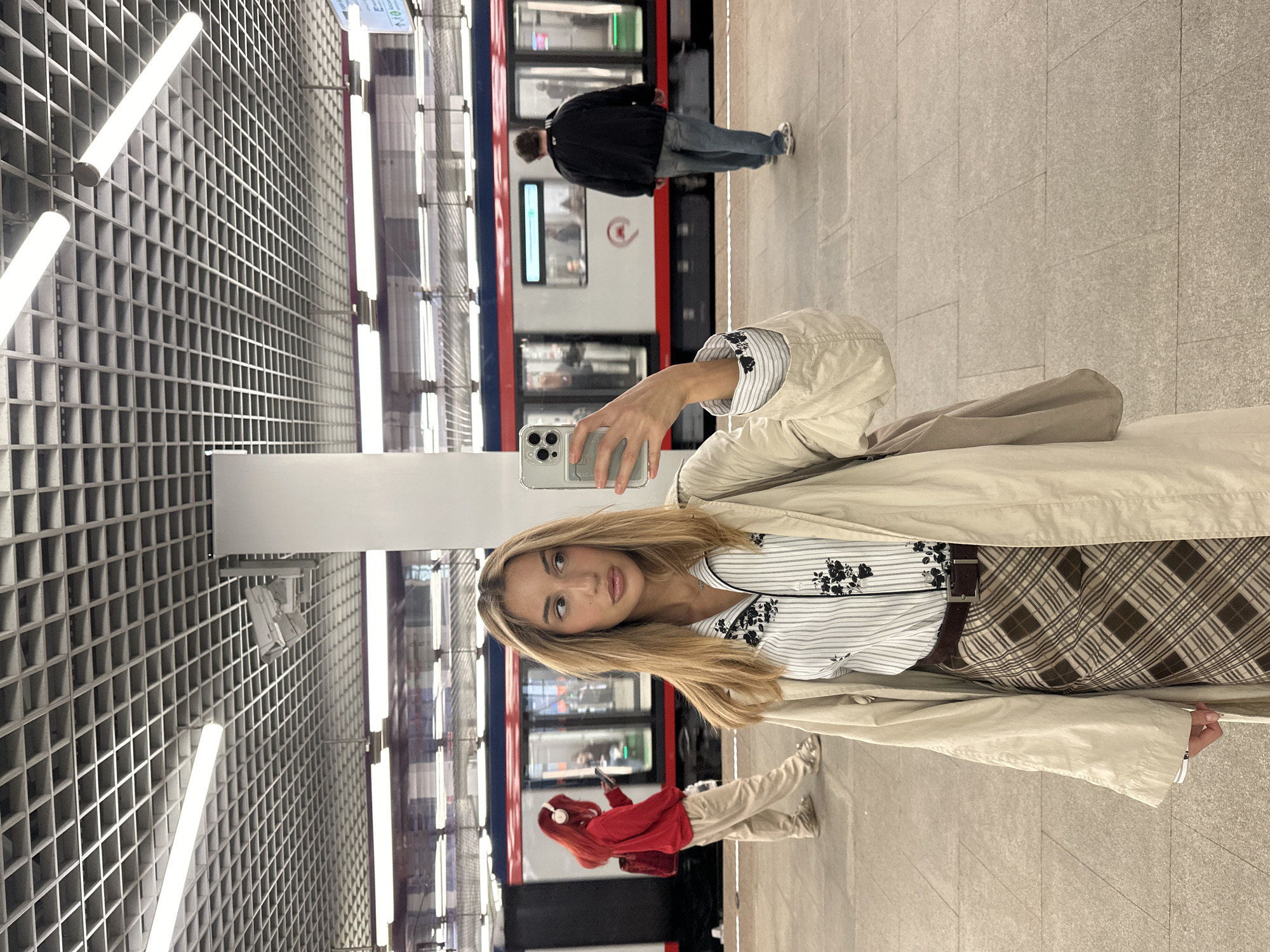 The media file captures a vertical, full-body selfie taken by a young woman on a Moscow subway platform. She is wearing a beige trench coat over a white and black striped shirt with floral embroidery, and a brown and beige plaid skirt with a dark brown belt. She holds a smartphone in her right hand, angled to take the picture.
The background shows a subway train with red and blue accents stopped at the platform. The platform itself is made of stone tiles. To the left, the station features a modern, grid-like ceiling with fluorescent lighting. In the background, another person in a black jacket and jeans stands near the train, looking away from the camera. Another individual with red hair and headphones is visible near the train doors.
The lighting suggests an indoor environment, typical of a subway station, without any clear indication of time of day or weather. The overall mood appears casual and everyday. There is no visible text in the image.