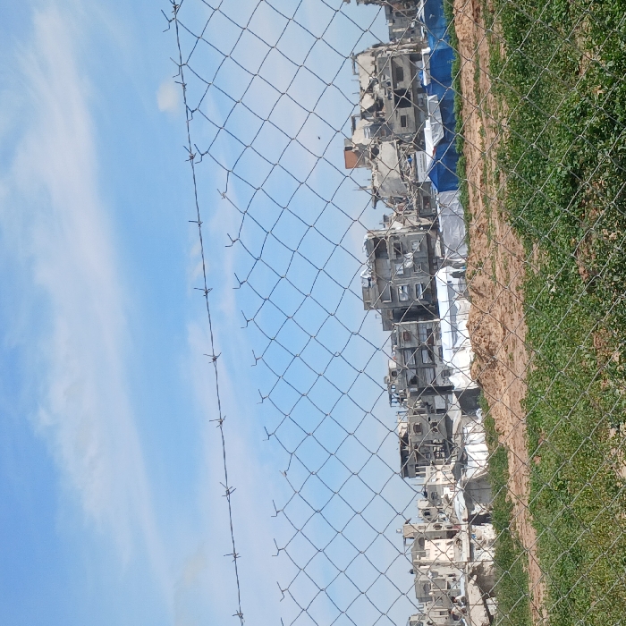 The image depicts a scene in Jabalia, Palestinian Territory, characterized by extensive structural damage and temporary habitation. In the foreground, a chain-link fence with a single strand of barbed wire running horizontally across its top edge spans the width of the frame. Below the fence, green vegetation, likely grass, transitions into an area of reddish-brown, disturbed earth. The mid-ground features several temporary shelters, including at least six white rectangular tents and two blue tents, erected on this ground amidst scattered debris. One dark-clad individual is observed walking from left to right in the lower-left mid-ground, near a white tent and a pile of light-colored rubble. In the background, a dense cluster of multi-story concrete buildings stands in various states of severe destruction, exhibiting missing walls, exposed rebar, and collapsed sections. The sky above is light blue with scattered white clouds.