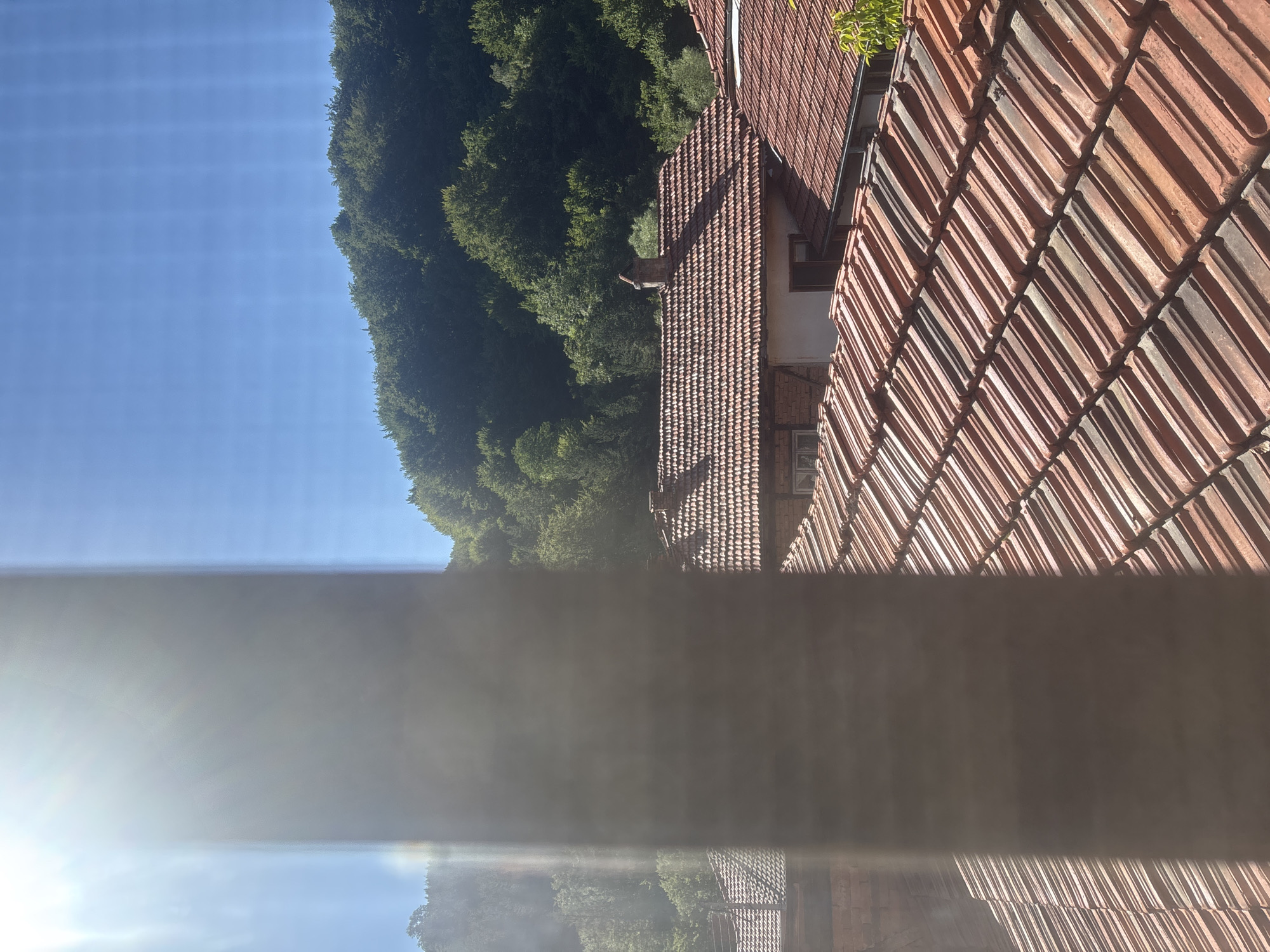 A bright sun shines from the upper left corner, casting lens flare across the image. A vertical, blurry pole or pillar obscures the center of the frame. Beyond the pillar, the foreground is dominated by an expanse of reddish-brown tiled roofs, characteristic of traditional Bulgarian architecture. In the middle ground, more tiled roofs are visible, some with brick chimneys. Behind the houses, a dense, green forest covers a sloping hill, leading up to a clear blue sky. The image appears to be taken from an elevated vantage point, possibly a window or balcony, looking out over a village in Shipkovo, Bulgaria.