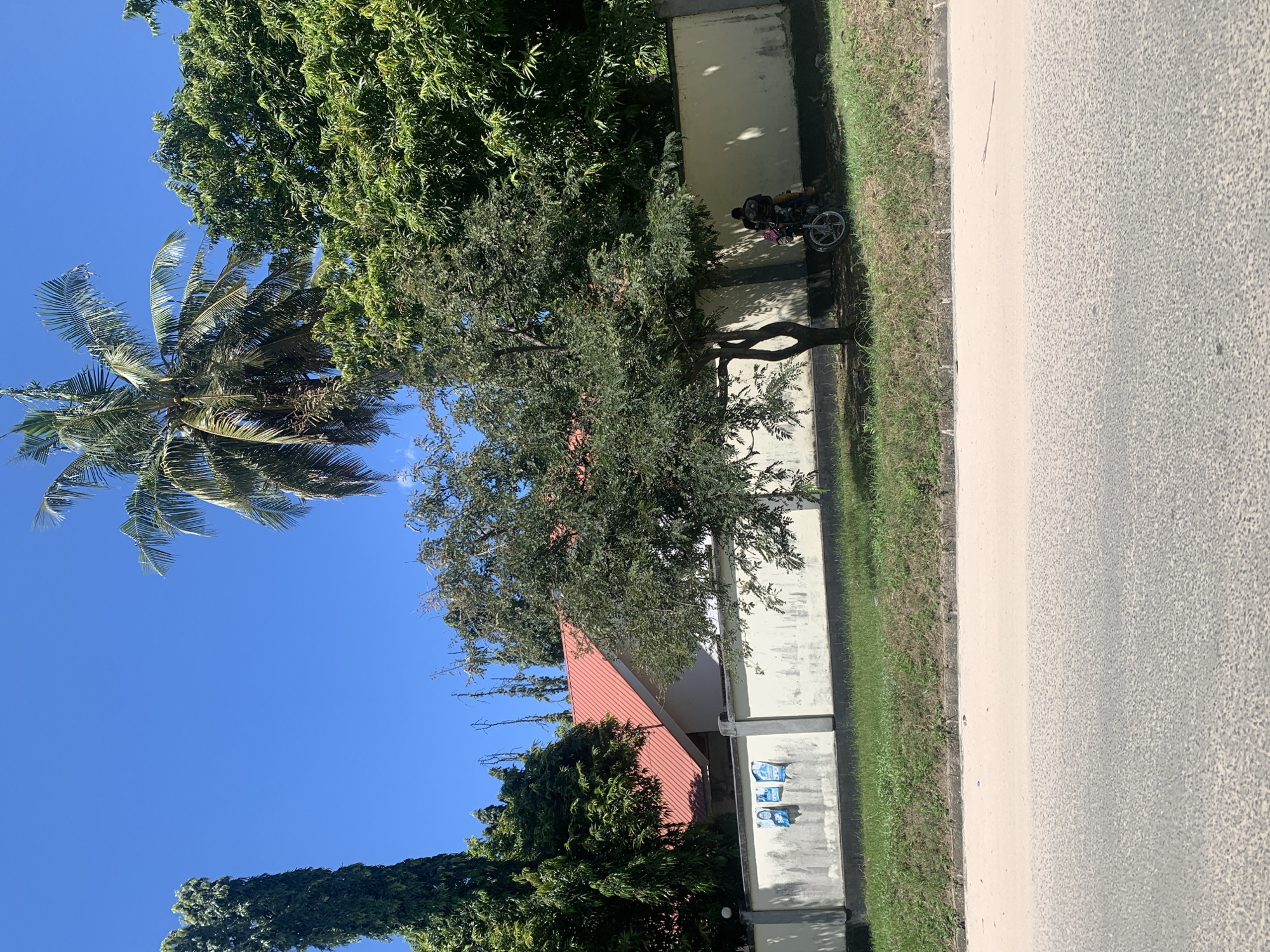 The image depicts a street scene in Dar es-Salaam, Tanzania. A white wall runs along the right side, bordered by a strip of green grass. A paved road occupies the immediate foreground. Beyond the wall, a building with a red roof is visible. Several trees, including a palm tree, are also in view. Parked near the wall, partially obscured by a tree, is a motorcycle. The sky is clear and blue.