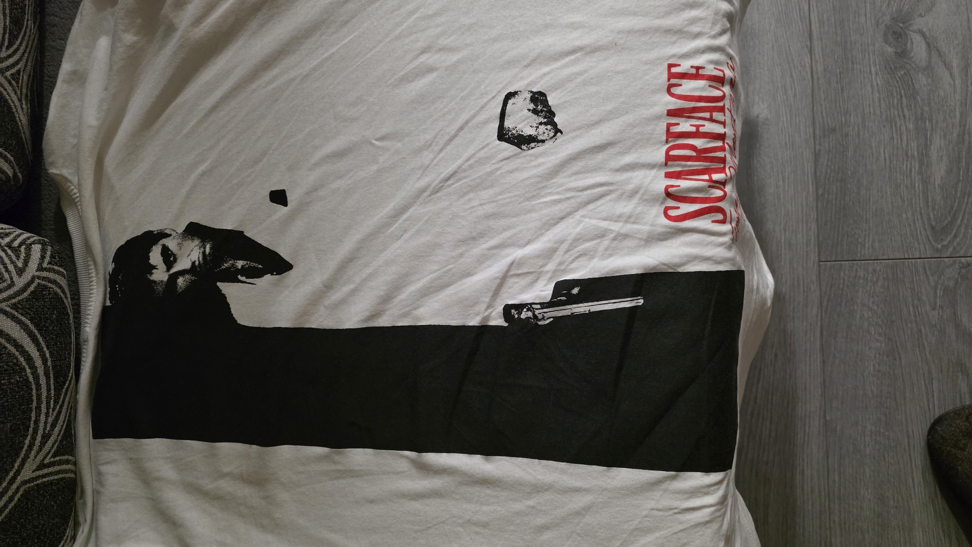 The image shows a white t-shirt. The t-shirt has a graphic print on it. The graphic print includes a depiction of Al Pacino in the movie Scarface, a gun, and the word "Scarface" in red lettering. The t-shirt is lying on a surface, possibly a couch or the floor. The background includes a wooden floor and a patterned couch. The exact location in Bogomilovo, Bulgaria is unknown.