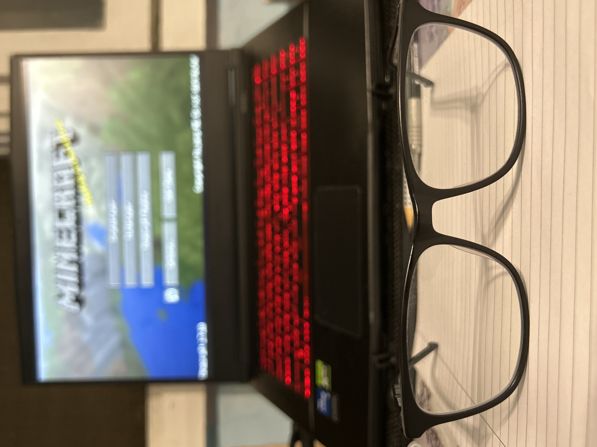 A laptop displays the Minecraft title screen with a red illuminated keyboard. A pair of black-framed eyeglasses sits on top of the laptop and a lined notepad. The location is unknown.
