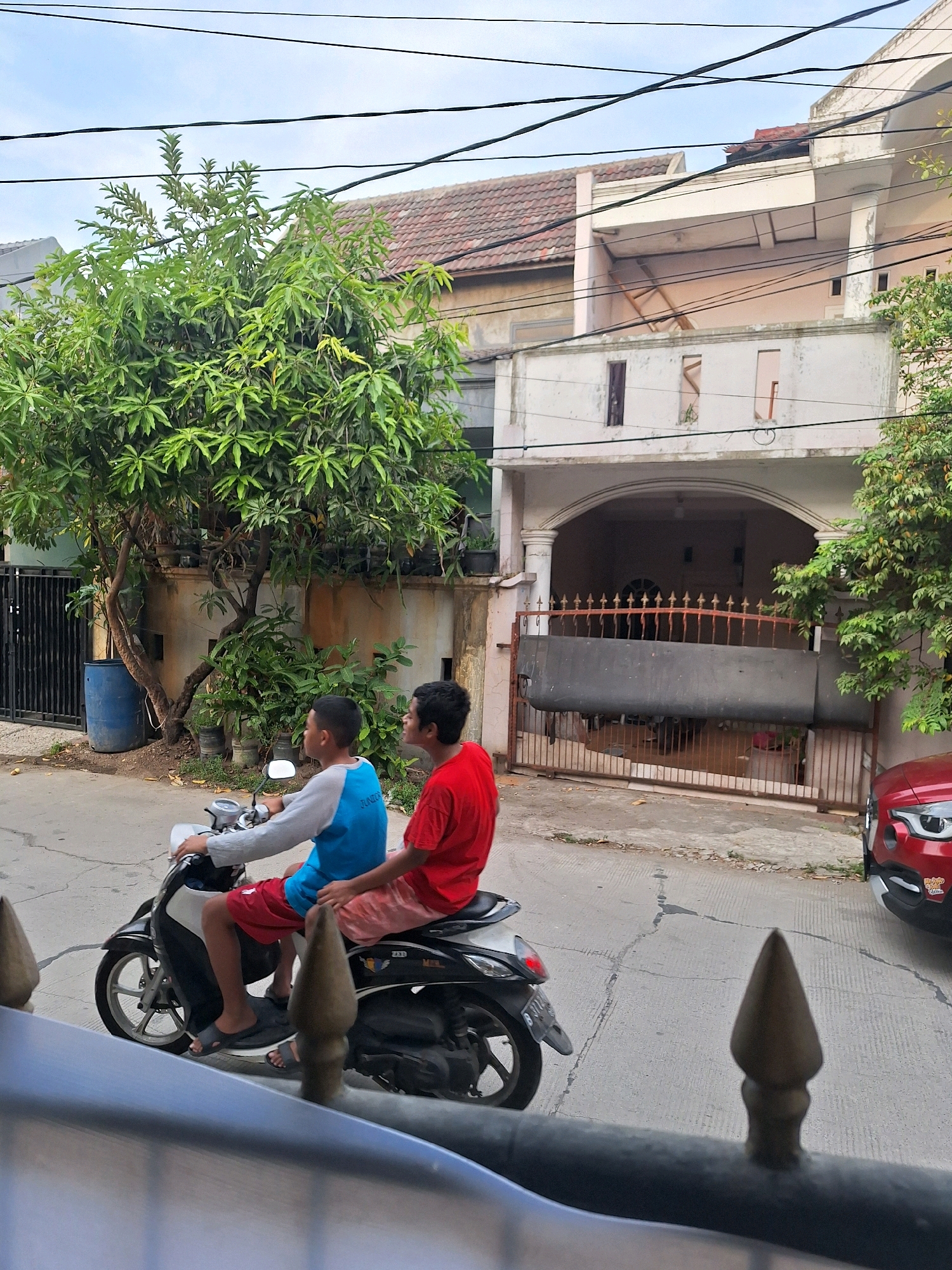Two young males are riding a scooter on a paved street in Setiamulya, Indonesia. The male in the front is wearing a blue long-sleeved shirt and red shorts, and the male in the rear is wearing a red t-shirt and pink shorts. They are both wearing sandals. A tree with green leaves is on the left side of the image, and a fence is behind the tree. Houses with red tile roofs are visible in the background. A red car is parked on the right side of the street. The sky is cloudy.