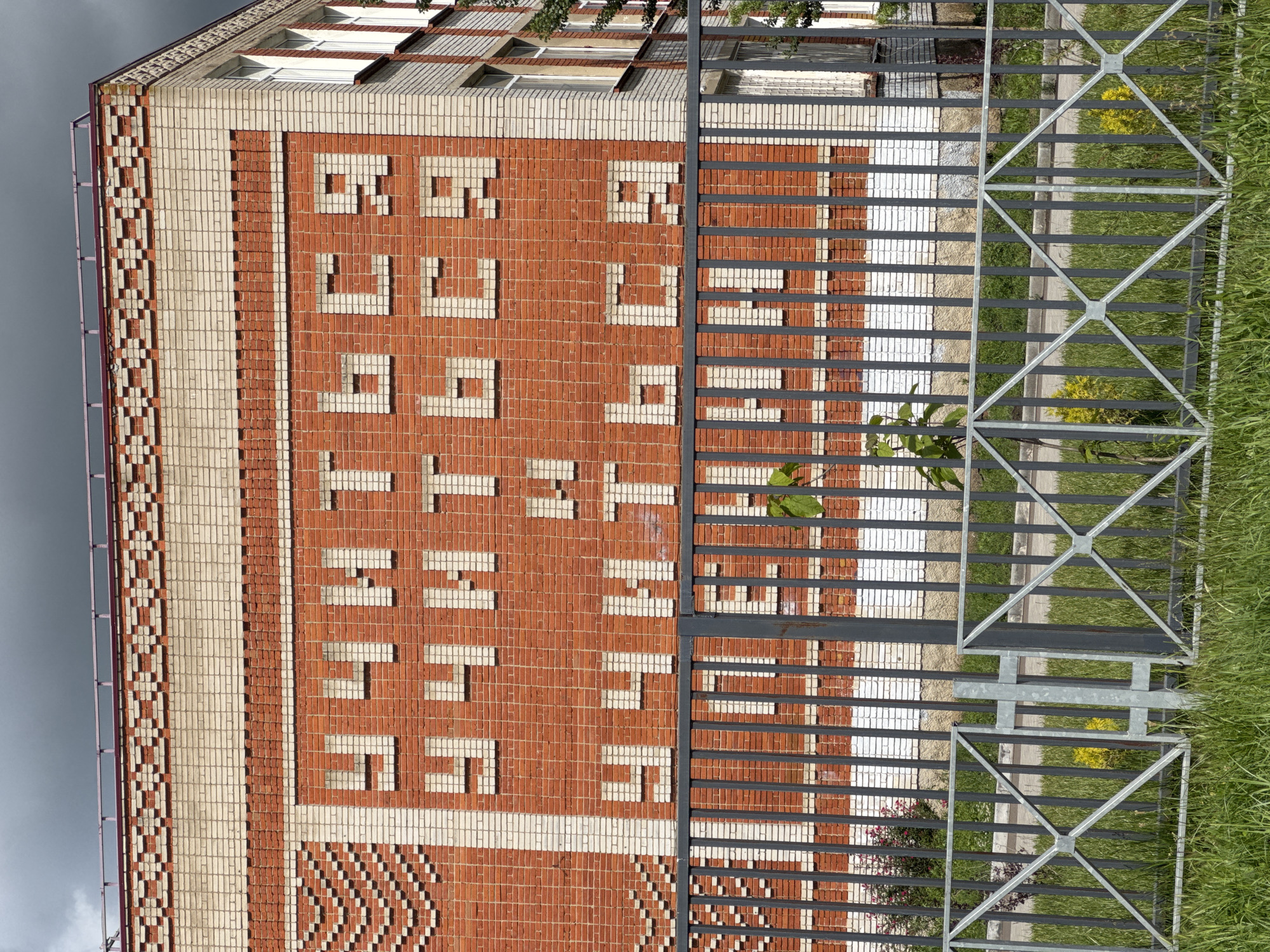 A building facade in Yelizavetinskaya, Russia, features a large inscription in Cyrillic script formed by contrasting light-colored bricks within a red brick wall. The inscription reads "ДЕТСКИЙ САД," which translates to "Kindergarten". To the right of the building, a metal fence encloses a grassy area. The sky is overcast. The building is constructed from bricks of different colors. There is greenery growing next to the fence.