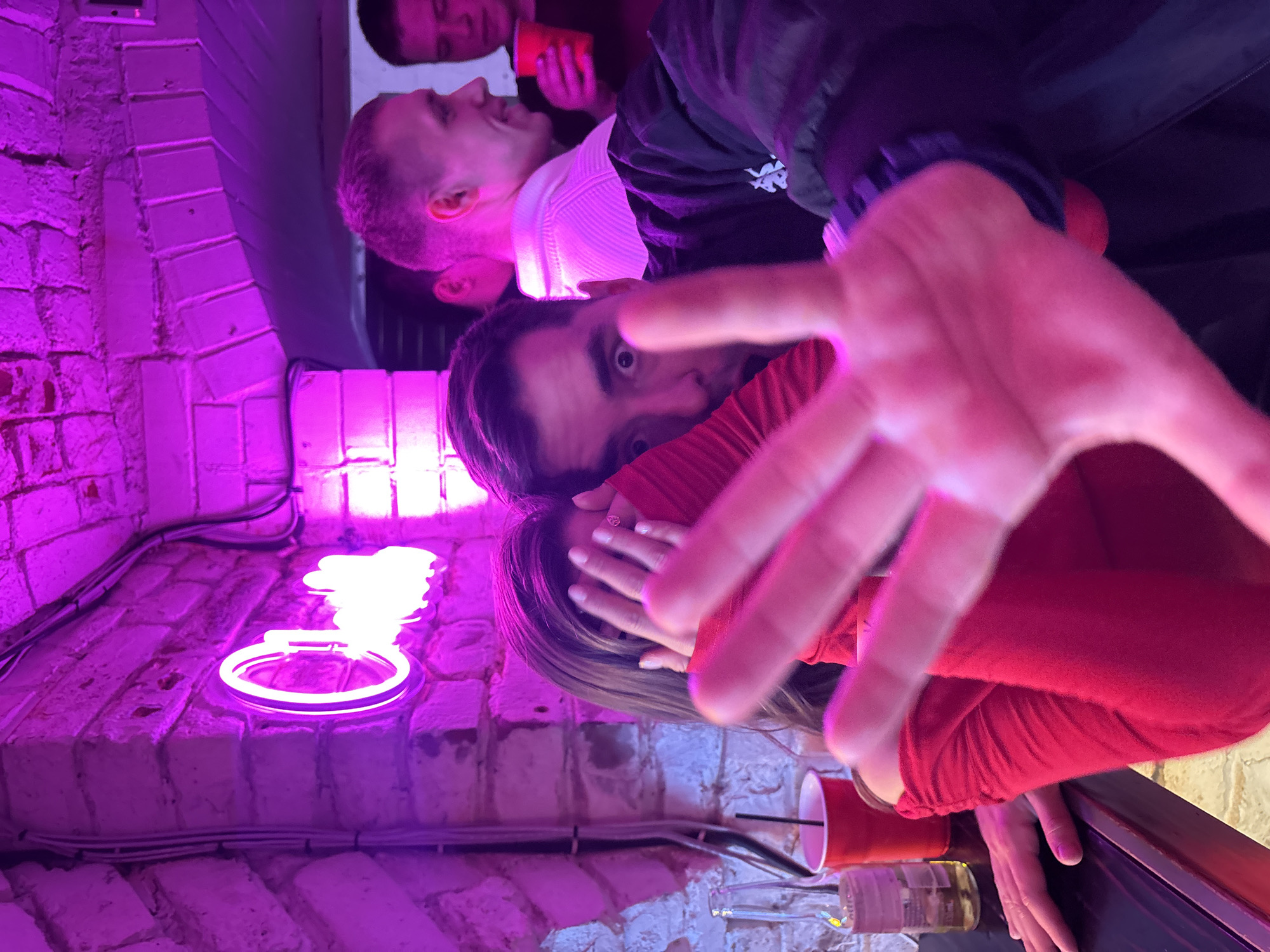 This image captures a vibrant, rotated scene from an indoor social gathering, likely in a bar or club in Krasnoyarsk, Russia. The setting is dominated by strong purple and pink neon lighting, casting colorful hues across the brick walls and the subjects.
In the immediate foreground, a hand with spread fingers reaches towards the camera, creating a dynamic, interactive feel. This hand belongs to a person wearing a red long-sleeved top, whose face is partially obscured by another hand, revealing a ring on one finger. Behind this, a man with dark hair and a surprised or wide-eyed expression stares directly at the camera, also in a red top.
Further back, another man with short hair and a white collared shirt beneath a dark jacket is seen in profile, looking to his right. In the upper background, another individual is visible, holding a red plastic cup.
The background features a textured brick wall, brightly illuminated by the neon lights. A prominent circular neon light fixture is mounted on the wall, contributing to the establishment's ambiance. Other objects include red plastic cups and a clear glass bottle on a surface. The general activity suggests a lively party or casual social event occurring at night, emphasized by the artificial lighting and celebratory items. The overall emotion conveyed is one of spontaneous interaction and merriment. No visible text is clearly legible in the image.