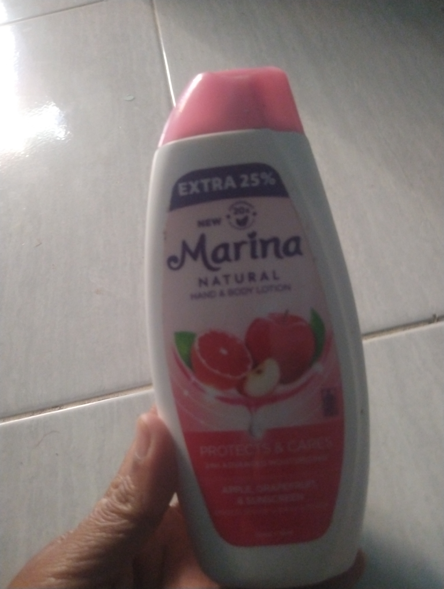 A hand holds a bottle of Marina Natural Hand & Body Lotion with an "Extra 25%" label. The bottle is white with a pink cap and features an illustration of an apple and grapefruit slices. Text on the bottle indicates it contains "Apple, Grapefruit, & Sunscreen". The background appears to be a tiled floor, consistent with indoor flooring or a paved outdoor area. The image is taken in Wanasaba Daya, Indonesia.