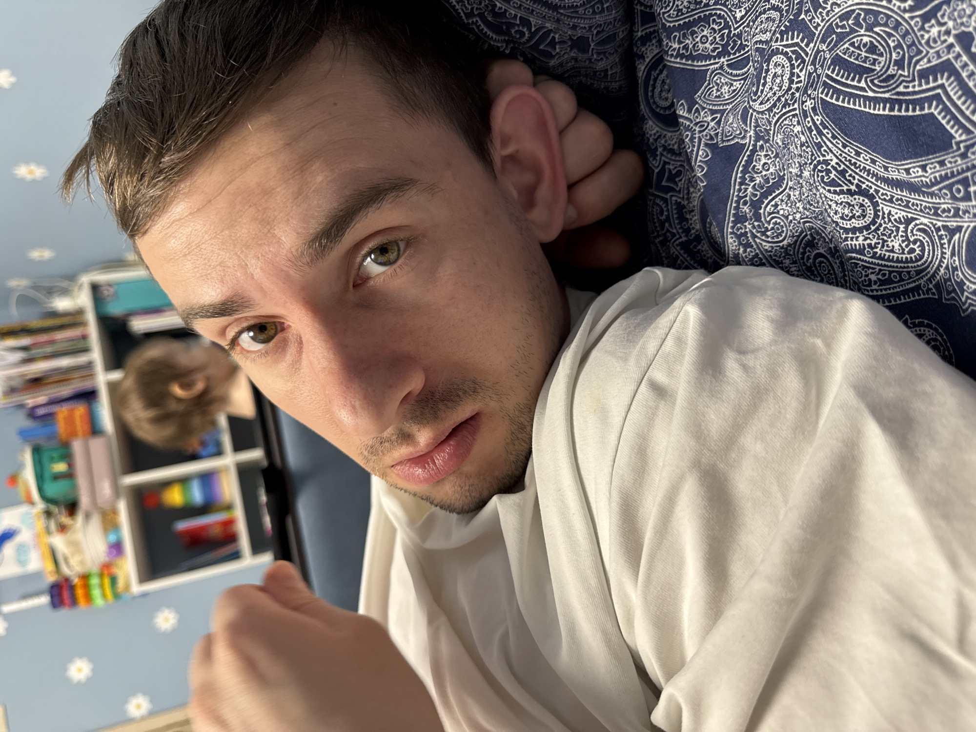 A close-up indoor selfie depicts a fair-skinned man with short dark hair, hazel eyes, and stubble. He is wearing a white t-shirt and appears to be lying down. The background includes a blue wall decorated with white flower patterns and a white shelving unit filled with various colorful objects. A blurry figure is visible near the shelving unit. The presence of the blue wall with flower patterns suggests the location is not Moscow. A blue paisley-patterned textile is seen behind the man's head. The man's right hand is visible in the lower left corner. He looks directly at the camera.