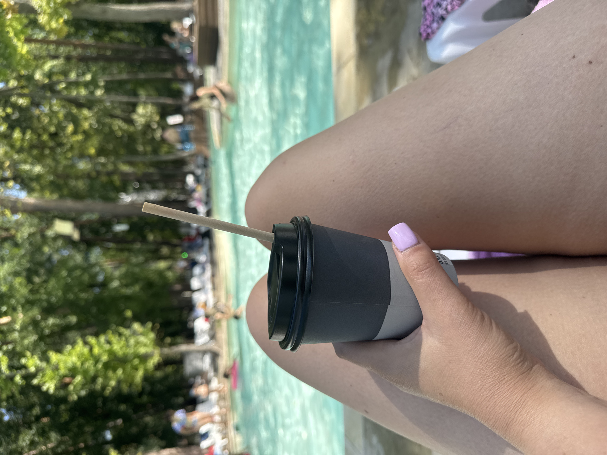 The image shows a woman's legs and hand holding a grey paper cup with a straw, resting near a swimming pool. In the background, there are trees and people swimming in the pool. The location is specified as Orlivshchyna, Ukraine, suggesting this is a resort or public pool facility. The lighting indicates it is daytime, likely sunny. The woman has light-colored skin and painted purple fingernails. The focus is on the foreground subject and her beverage, with the background providing context of an outdoor recreational setting.
