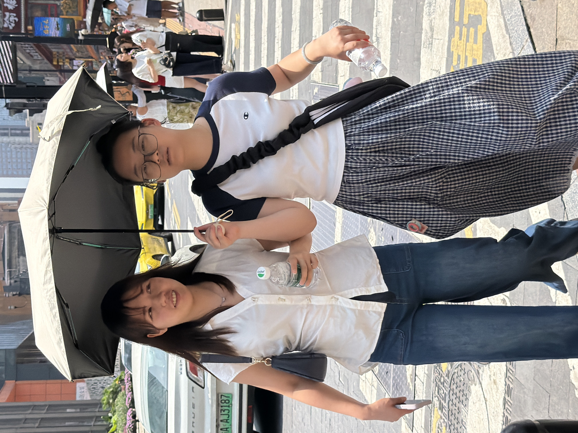 Two young East Asian women walk together under a large black umbrella on a pedestrian crosswalk in Yuzhong District, China. The woman on the left, with shoulder-length dark hair and bangs, wears a white short-sleeved button-up shirt and blue denim wide-leg pants. She carries a small dark purse on her left shoulder and holds a clear plastic water bottle in her right hand. The woman on the right, wearing glasses and her hair pulled back, is dressed in a white t-shirt with navy raglan sleeves and a navy and white plaid skirt. She has a black crossbody bag with white stripes over her right shoulder and holds a clear plastic water bottle in her right hand. In the background, a white car with a license plate reading "A AJ3187" is visible, along with other pedestrians, buildings, and street signage. The environment appears to be a sunny urban street.