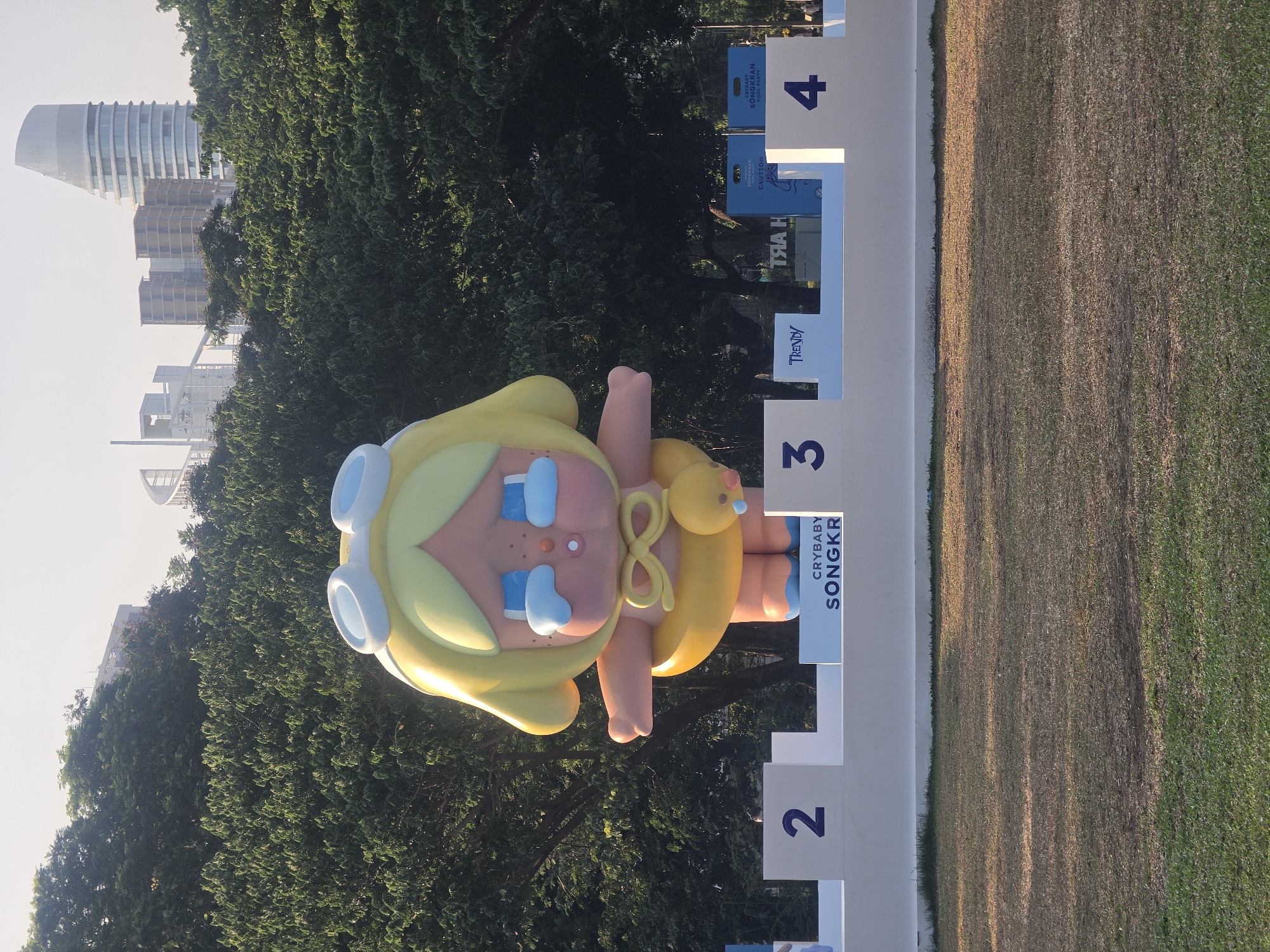This image, rotated 90 degrees, captures a large, inflatable cartoon character, likely "Crybaby" from Pop Mart, situated outdoors in an urban environment. The character, with blonde hair, light blue goggles, blue tearful eyes, blushed cheeks, and a yellow pacifier with a matching cord, is the central focus. It appears to be wearing a light blue diaper or briefs.
Behind the inflatable figure, a lush line of green trees partially conceals a modern city skyline. Several high-rise buildings are visible, including a distinctive white skyscraper with a curved, multi-tiered design, indicative of a contemporary metropolitan setting. To the right of the character, a white barrier or display structure is present, segmented with large dark blue numbers "2", "3", and "4". Blue panels attached to this structure display text such as "CRYBABY SONGKRAN", "Trendy", and "CAUTION", suggesting an event related to the Thai New Year festival.
The ground is a grassy field, showing variations in color from green to dry brown, implying an open park or event space. The lighting is soft and warm, suggesting either early morning or late afternoon, with clear weather conditions. The scene depicts a public art installation or display for a "Crybaby Songkran" event, likely located in Bangkok, Thailand.