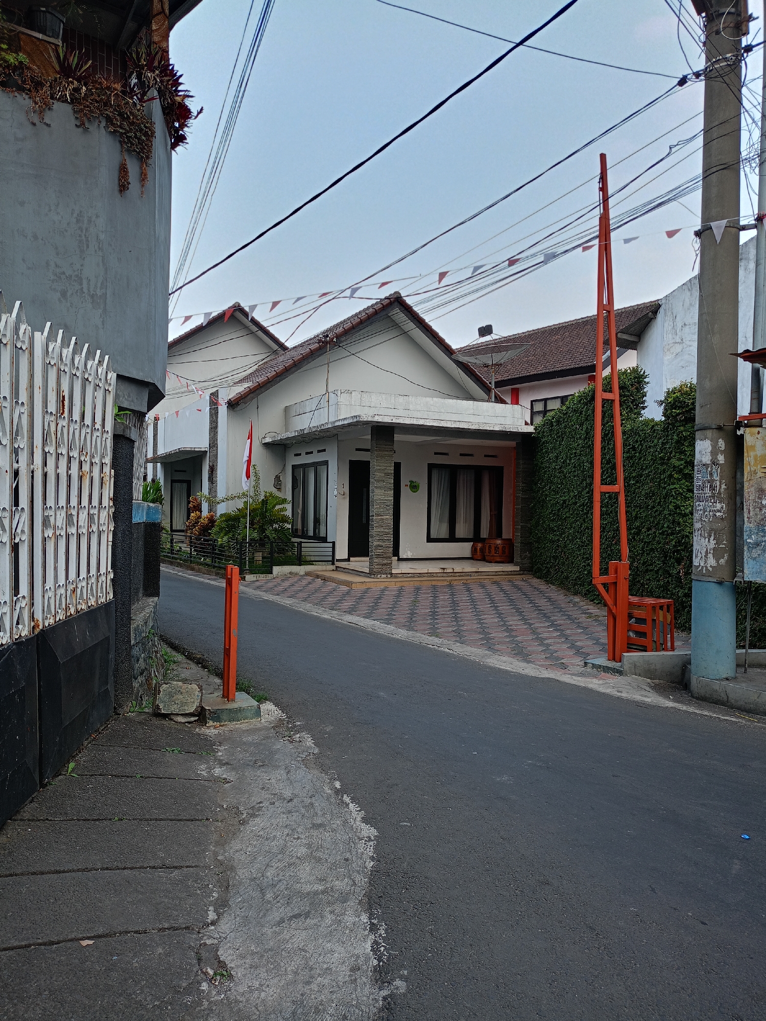 The image depicts a residential street in Batu, Indonesia, likely in the evening or early morning given the overcast sky and subdued lighting. A paved road curves through the scene, bordered by a sidewalk on the left and a driveway on the right. A white metal fence with decorative elements runs along the left side of the road, partially obscuring a grey wall with plants cascading from above.
On the right side of the road, a modern house with a white facade and dark-tiled roof is visible. The driveway leading to the house is paved with a repeating pattern of red and grey hexagonal tiles. A tall, bright orange barrier arm, commonly used for access control, stands prominently near the entrance to the driveway, positioned next to a thick green hedge. A small Indonesian flag, red and white, flutters in front of the house.
Further down the street, another similar house is partially visible, also featuring a tiled roof and white exterior. Overhead, a network of electrical wires stretches across the sky, punctuated by small red and white triangular flags strung between poles, suggesting a festive or celebratory occasion. The environment appears quiet and orderly, typical of a residential neighborhood. No people are present in the image, and no specific actions are being taken. The focus is on the architectural elements and the streetscape of this Indonesian city.