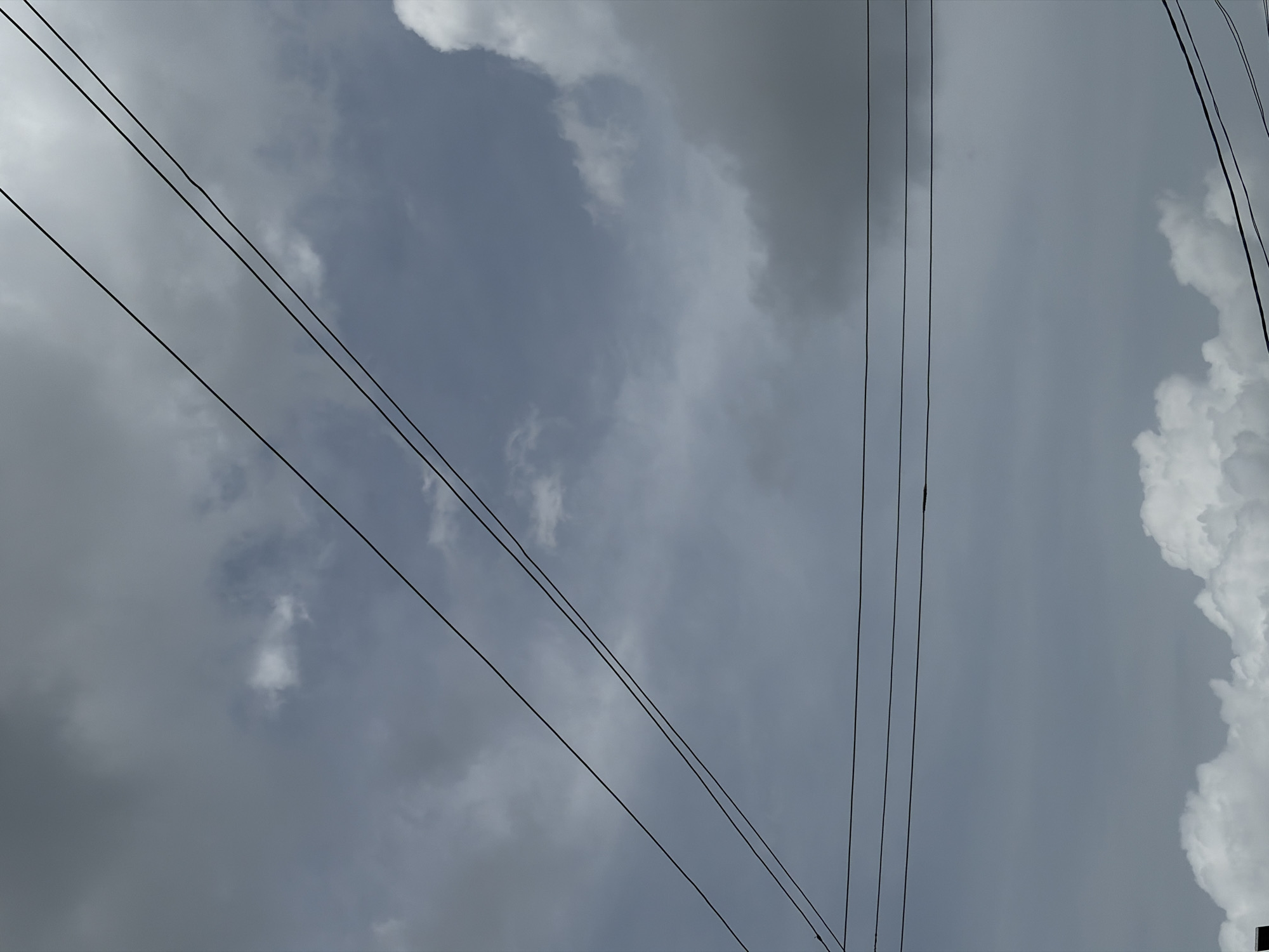 The image shows an upward view of a cloudy sky in Igboefon, Nigeria. Black power lines crisscross the frame. The clouds are predominantly gray with some patches of white.