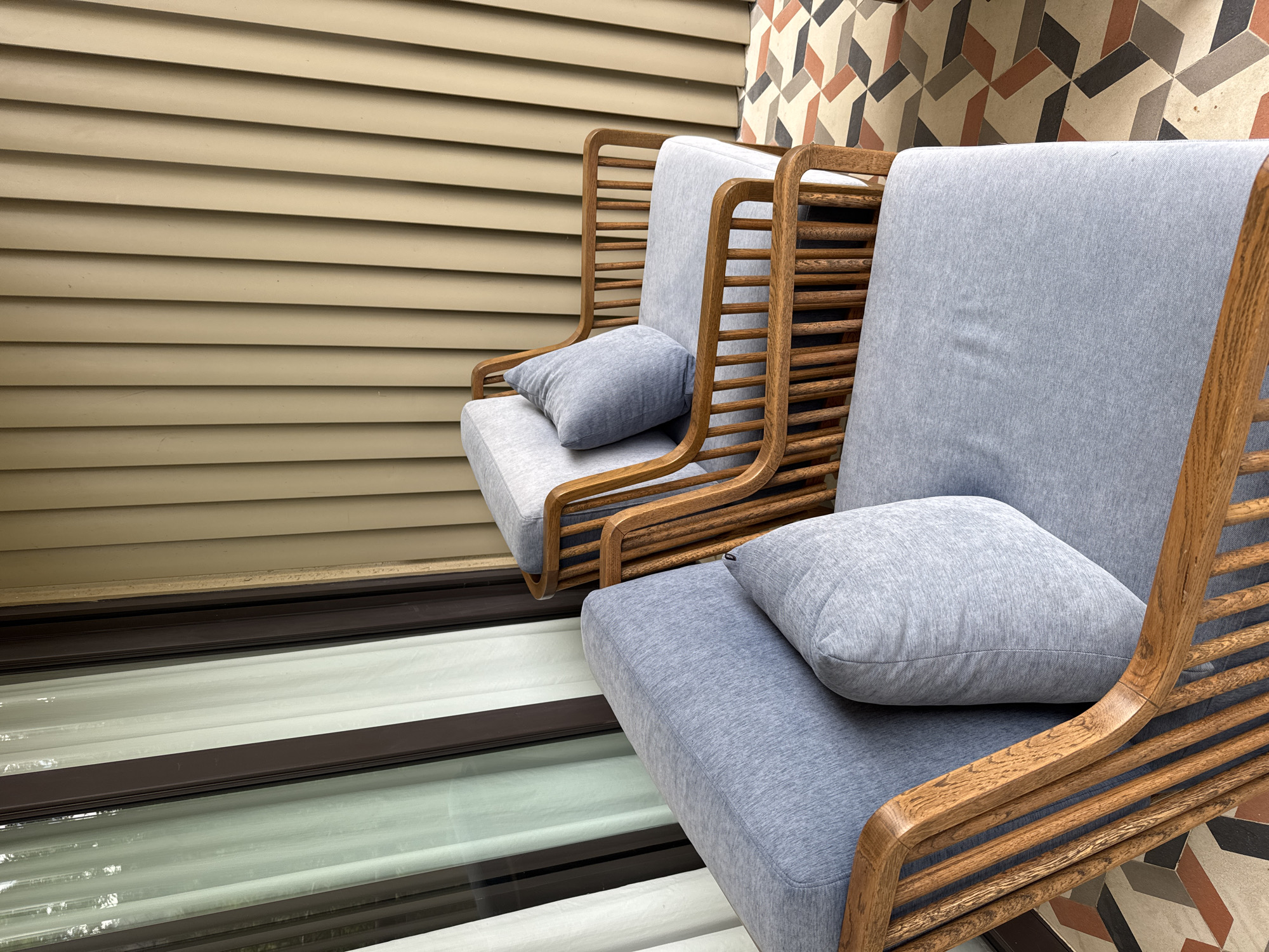 Two light blue cushioned armchairs with natural wooden frames and slatted details are positioned on a geometrically tiled patio in Púng Luông, Vietnam. The chair in the foreground, partially visible on the left, features a rectangular throw pillow. Behind it, a second, more completely visible chair mirrors its design. To the left, a dark-framed glass door or window with sheer white curtains reflects ambient light. To the right, a textured wall with vertical panels in a beige hue extends upwards. The floor features a multi-colored geometric tile pattern in shades of cream, terracotta, dark gray, and light gray. The scene is illuminated by natural daylight. No individuals are present.