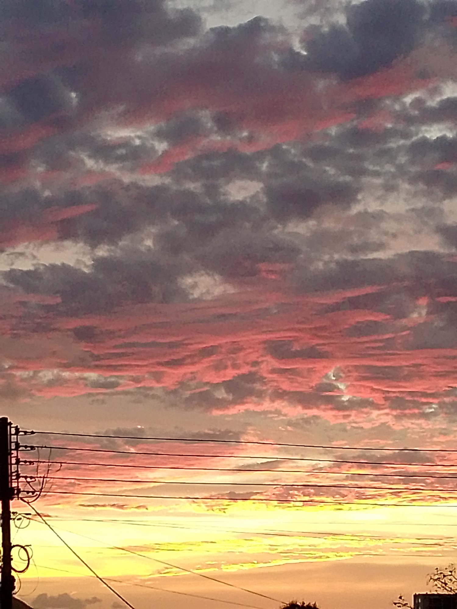 The image shows a vibrant sunset sky above Juan Griego, Venezuela. The sky is filled with long, streaky clouds colored in shades of pink, gray, and white. Below the clouds, a band of yellow and orange light from the setting sun illuminates the horizon. In the foreground, a utility pole with wires stretches across the scene. The lower portion of the image contains outlines of rooftops and foliage.