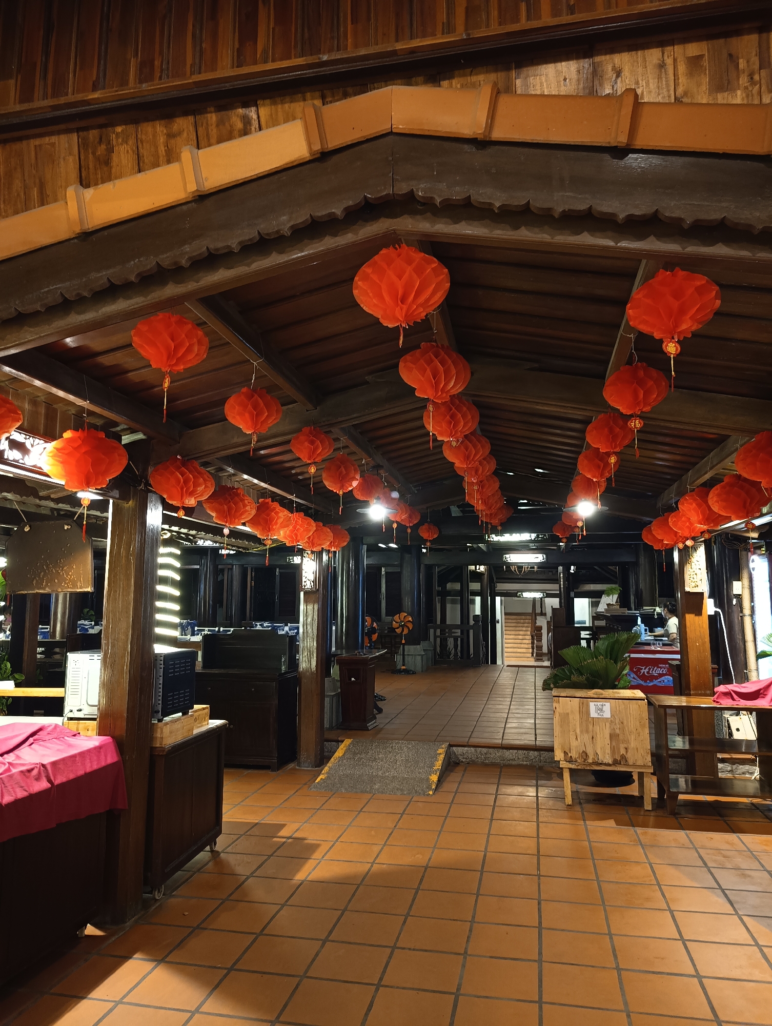 This is a photograph of an indoor restaurant or dining area, likely in Phu Thuy Ward, Vietnam, decorated with numerous traditional red paper lanterns. The lanterns are suspended from the wooden ceiling beams, casting a warm, festive glow throughout the space. The architecture features exposed wooden beams and paneling, creating a rustic and traditional ambiance.
The scene appears to be set during the evening or night, as indicated by the artificial lighting and the subdued natural light filtering in. The floor is tiled with large, square terracotta tiles.
Various objects are visible in the foreground and background, suggesting a functioning establishment. To the left, there's a table covered with a red cloth. Further back, several wooden cabinets and what appear to be kitchen appliances or storage units are arranged. In the middle ground, a wooden staircase leads upwards. To the right, a counter area is visible with a person behind it, possibly a staff member. A wooden crate with some greenery is also present.
The red lanterns are the most prominent decorative elements, evoking a sense of celebration or cultural tradition. There is no visible indication of the weather. The overall mood is inviting and festive, likely a preparation for or during a special occasion. Some of the lanterns have small golden charms attached. One visible sign reads "Hilace".