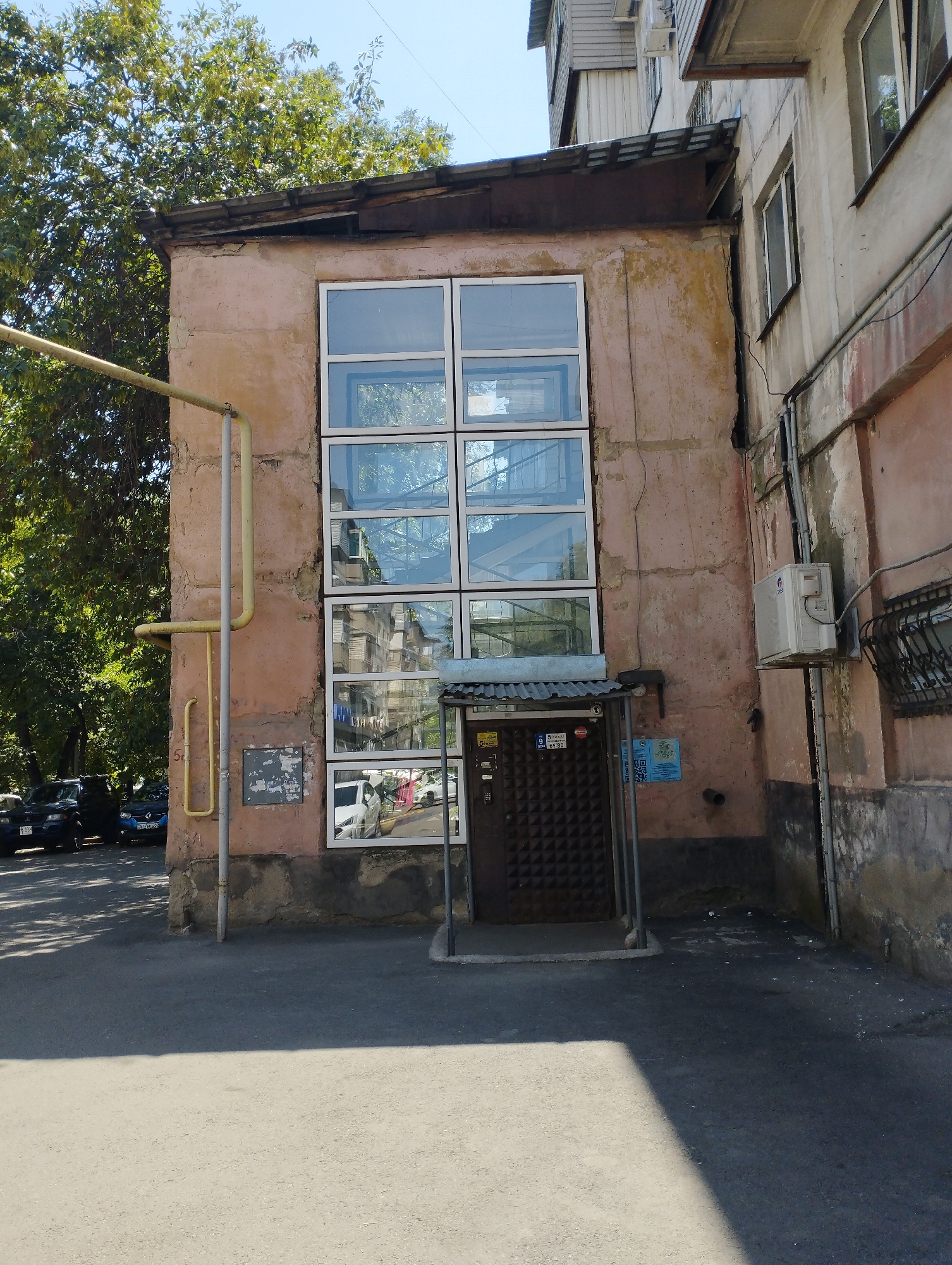 The image captures an exterior view of a dilapidated building, likely in the city of Almaty, Kazakhstan. The building features a large, multi-pane window dominating its facade, reflecting the surrounding environment. To the left of the window, a yellow gas pipe is visible, attached to the textured, pale pink wall. A dark brown metal door is situated beneath the window, protected by a corrugated metal awning. Signs with Cyrillic lettering are posted near the door. The ground in front of the building is asphalt. In the background, lush green trees are prominent, and several cars are parked along the street. The overall impression is one of urban decay, with the building showing signs of age and neglect. The sky above is clear and blue.