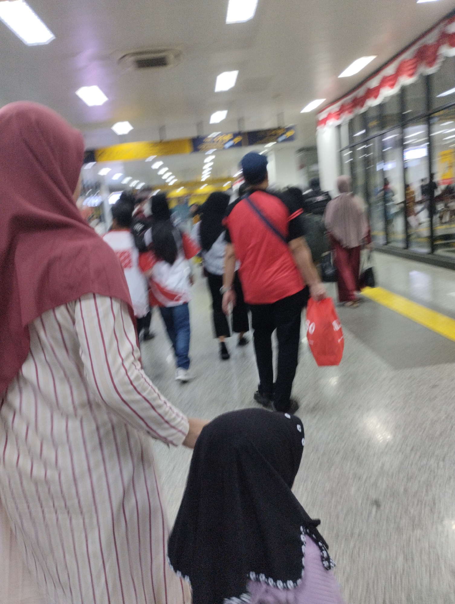 The image depicts a scene inside a building with a high ceiling containing fluorescent lights and what appears to be an air conditioning unit. In the foreground, the back of a person wearing a maroon hijab and a striped dress is visible. To the right of this person, another person, possibly a child, is wearing a black hijab and a purple garment.
In the middle ground, a group of people are walking. One individual in a red shirt and black pants is holding a red plastic bag. Other individuals are seen in white shirts with red accents and blue jeans.
The background shows large glass windows with red and white decorative banners hung above them. The floor is tiled with a yellow stripe along the edge of a pathway. The overall impression is of a public indoor space, possibly a transportation hub or a large waiting area. The presence of the banners and the general atmosphere suggest a festive or organized event. Without further context, it is not possible to definitively identify the specific location within South Jakarta, Indonesia, but it could be an area within a large station or complex.