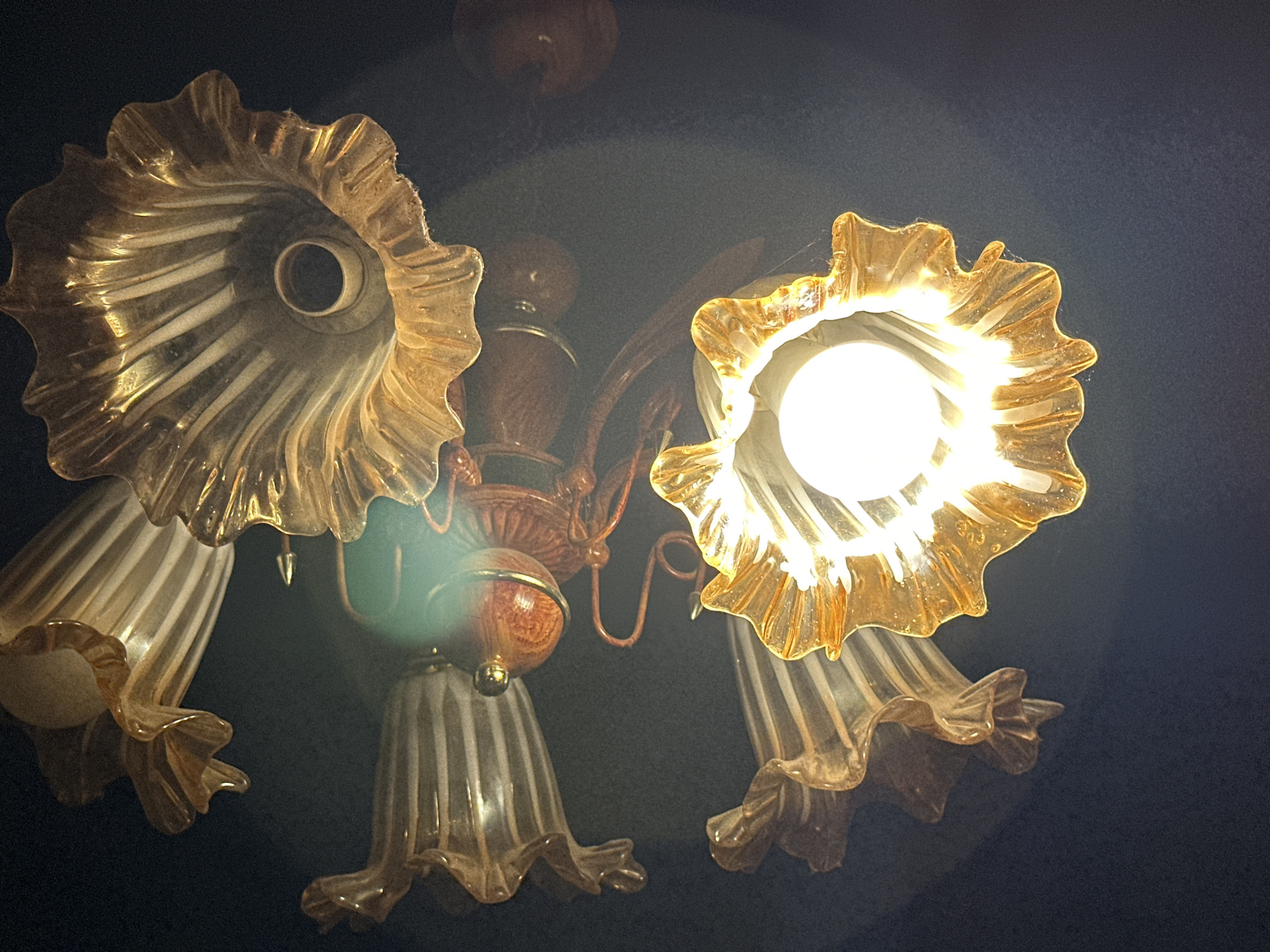 The image shows a close-up view of a chandelier with amber-colored glass shades, one of which is illuminated by a bright white light bulb. The chandelier has a metallic, possibly brass, frame and decorative elements. Several of the glass shades are flared and ribbed, with ruffled edges. The background is dark, suggesting the photo was taken in low light conditions. There are no people or animals visible in the image. The location is identified as Talas City, Kyrgyzstan, however, the image itself does not contain any visual cues to confirm this location; it depicts an indoor lighting fixture.