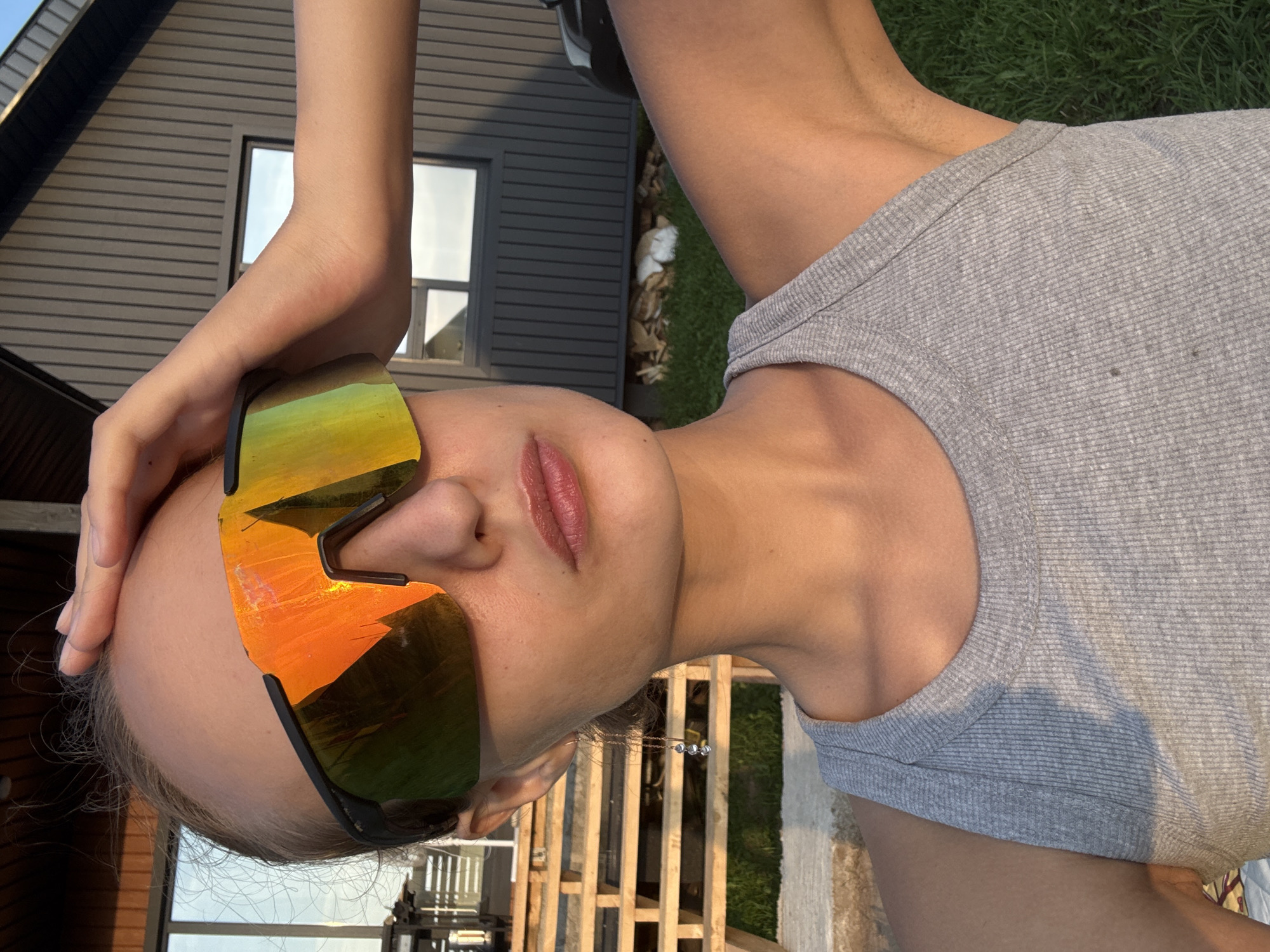 The image shows a close-up of a person's face, turned sideways. They are wearing large, reflective sunglasses with orange and green lenses, and a grey ribbed tank top. Their left hand is resting on their forehead. The background appears to be outdoors, with a modern-style house with dark siding and a window visible to the left. Behind the person, there is a wooden fence or structure, green grass, and some fallen leaves. The lighting suggests it is daytime, possibly late afternoon or early evening, given the warm tones reflecting on the sunglasses. The overall mood seems relaxed and casual, as if the person is enjoying a sunny day outdoors. No text is visible in the image. The location context is Podshivalovo, Russia.
