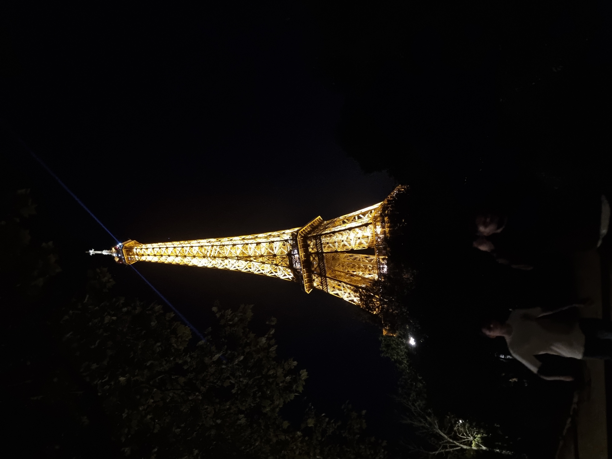 The image depicts the Eiffel Tower at night, illuminated with golden lights. A blue beam shines from the top of the tower. There are trees visible in the foreground, partially obscuring the lower part of the tower. Two people are visible in the bottom right corner. The location is Paris, France.