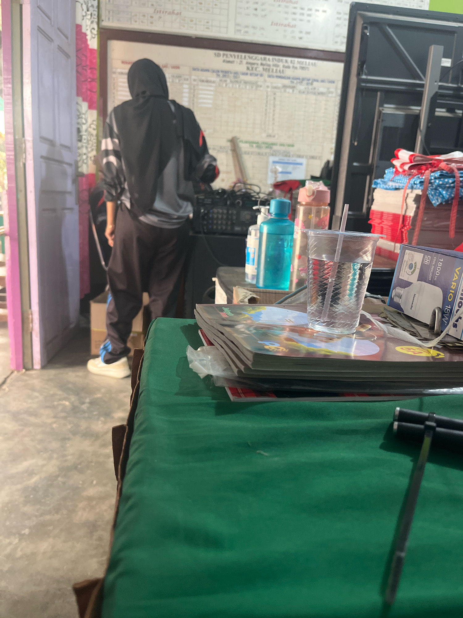 In Meliau Hulu, Indonesia, an individual wearing a dark hooded jacket and dark pants is standing in front of a bulletin board. The bulletin board displays charts and schedules. To the right of the individual, there is audio equipment and a stack of items that appear to be books or magazines. In the foreground, a glass of water with a straw sits on top of a stack of magazines, and a green cloth covers a surface. Further to the right, a lightbulb box is visible. The scene appears to be indoors, possibly in a school or community building.