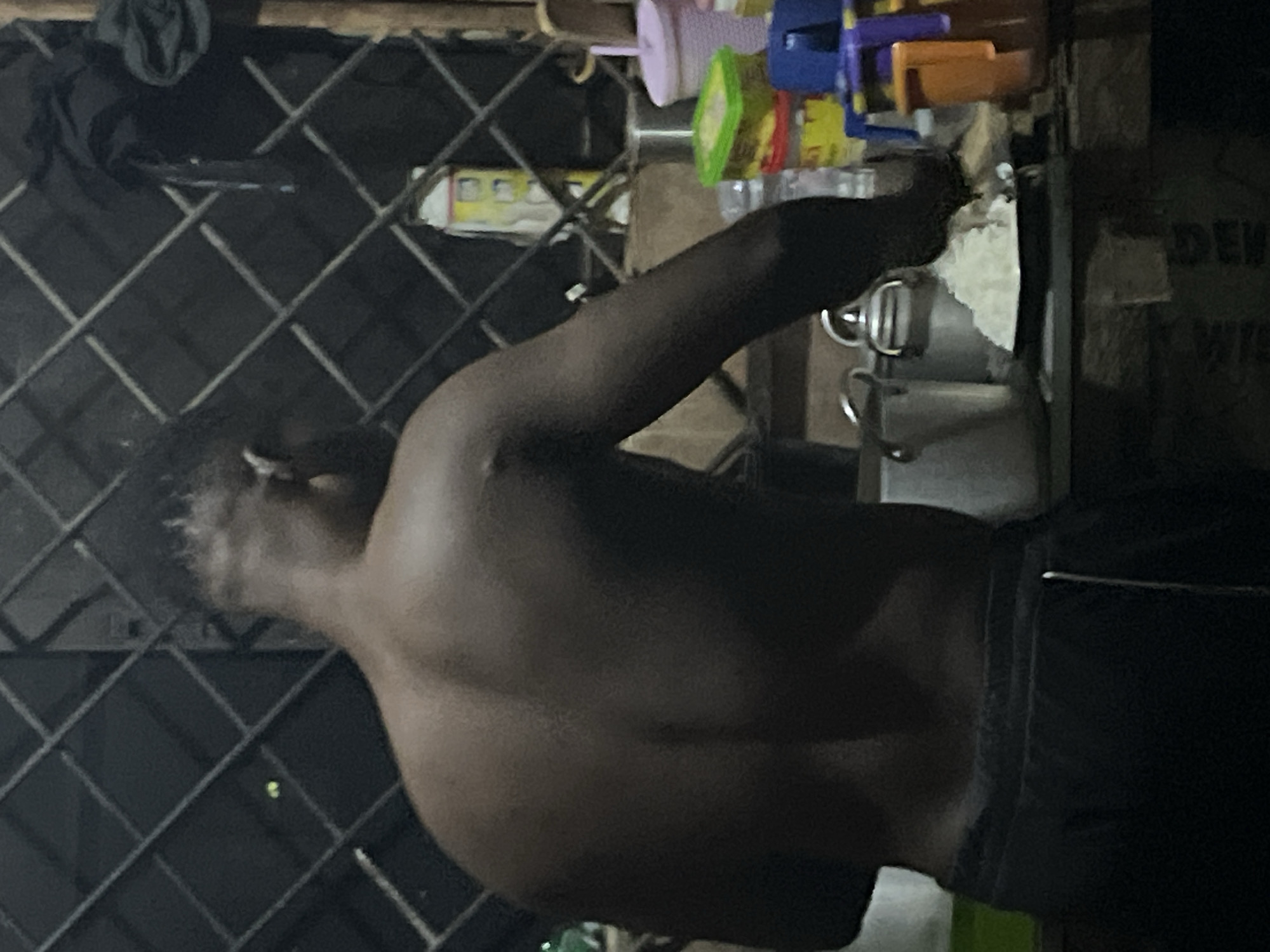 The image shows the back of a person, appearing to be a man, from the shoulders down to the waist, wearing black shorts. He has dark skin and short, dark hair. He is standing in front of a metal grate that is partially obscured by his body. Behind him, there are various objects on a shelf, including a white bottle, a pink cup, and a green and yellow container. To the right, there is a blue and orange object that resembles a toy or storage container. In the foreground, on the shelf, there is a plate of white rice next to a large metal pot with handles. The scene appears to be indoors, with a dimly lit environment and a rough wooden surface beneath the shelf.