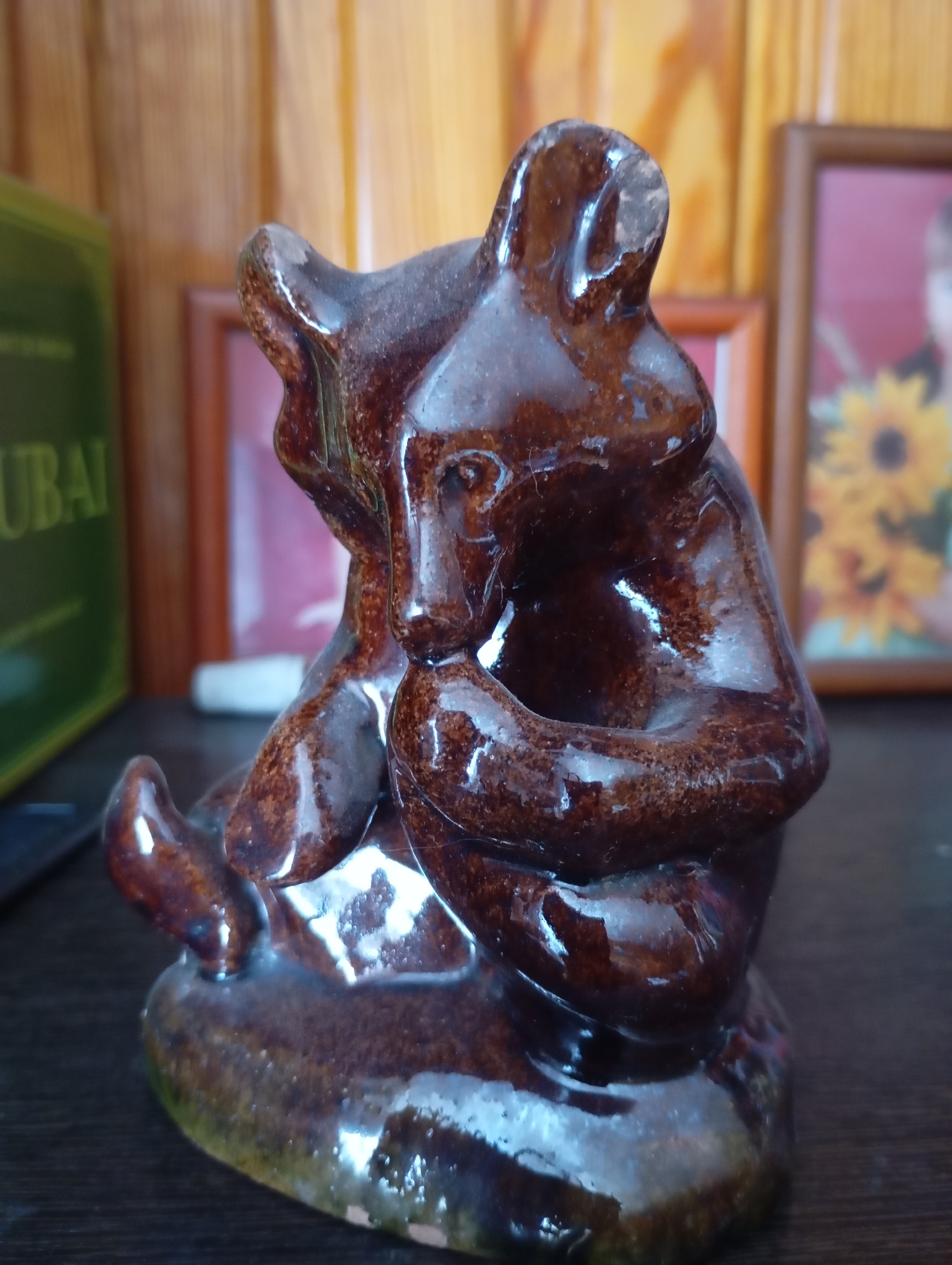A close-up, slightly low-angle shot captures a brown ceramic figurine of a bear cub seated with its paws clasped. The figurine has a glossy finish and appears to be resting on a dark surface, possibly a table. In the background, a wooden paneled wall is visible, along with two framed pictures. One frame on the left has a green book partially visible next to it with the word "UBAI" on its spine. The other frame on the right contains a picture of a person and a sunflower. The lighting suggests an indoor setting, likely a home environment in the village of Lnikha, Russia.