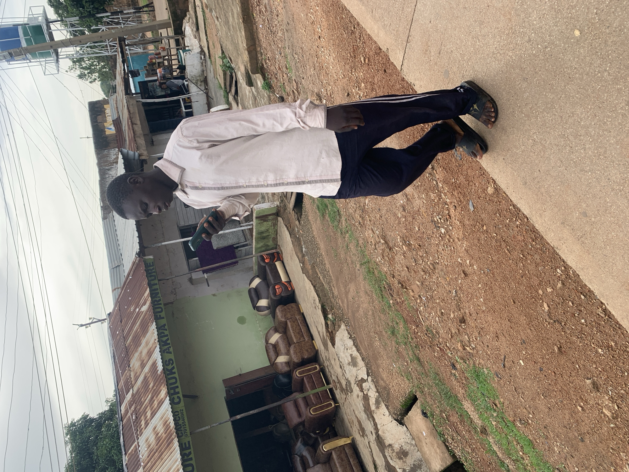 The image shows a young Black man standing outdoors on a paved walkway next to a dirt path. He is wearing a light-colored long-sleeved shirt and dark pants with white stripes down the sides. He is holding a dark-colored object, possibly a phone, in his right hand. He is wearing dark sandals on his feet.
Behind him, there are buildings, including a structure with a corrugated metal roof that appears weathered. A sign on this building reads "CHUKS AKPA FURNITURE" and includes a phone number. There are also several brown leather chairs or sofas displayed outside the building. Another building in the background has a light green facade.
The scene appears to be a commercial area, possibly a furniture shop, in a developing region. The ground is a mixture of concrete, dirt, and some sparse vegetation. The sky is overcast, suggesting a cloudy day. The orientation of the image is such that the subjects are nearly horizontal.