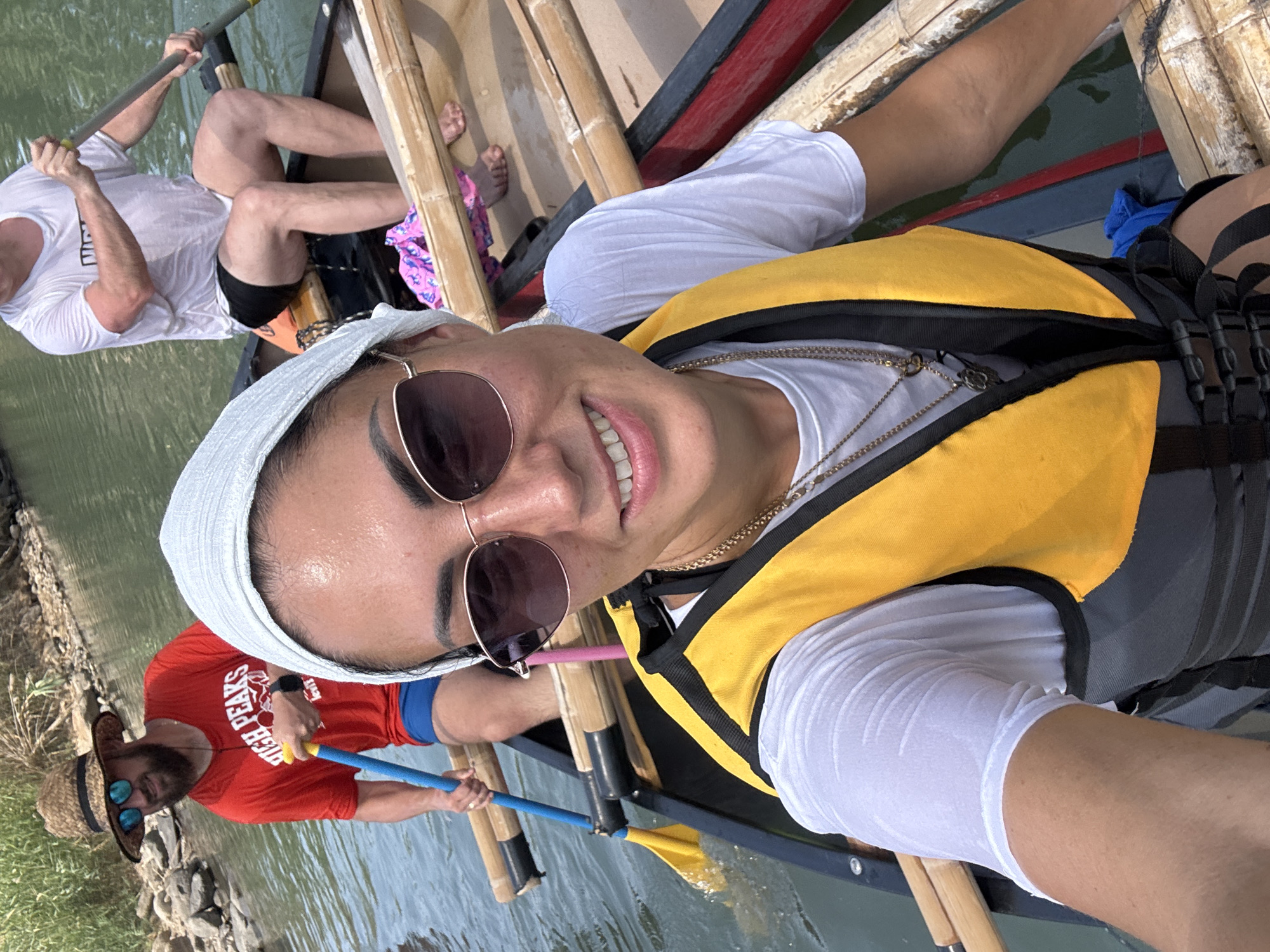 This image shows three people in a canoe on a body of water. The person in the foreground is a woman wearing sunglasses, a white headscarf, a white t-shirt, and a yellow life vest. She is smiling at the camera and appears to be taking a selfie. Behind her, to the left, a man wearing a straw hat, sunglasses, a red t-shirt, and blue shorts is paddling the canoe. He is holding a blue paddle with a yellow blade. To the right, another person, a man wearing a white t-shirt and black shorts, is also paddling. The canoe is made of wood and has bamboo railings. The water is green and there is vegetation on the bank in the background. The location is identified as a river in the Emek HaYarden Regional Council, Israel.