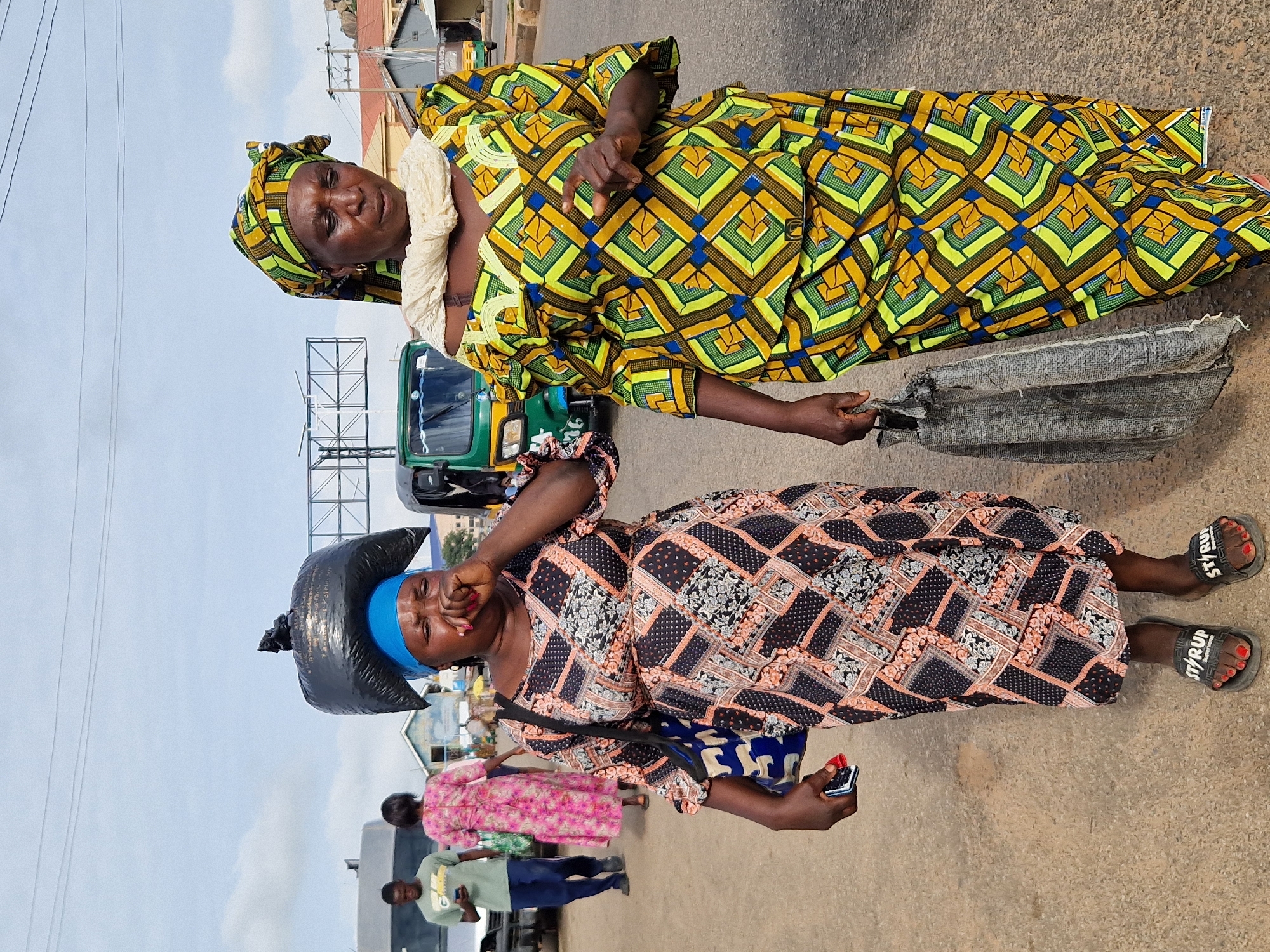 This is a medium shot, eye-level, outdoor photograph taken on a paved road in Kabong, Nigeria. The foreground is dominated by two women walking towards the viewer. The woman on the left, closer to the camera, is wearing a blue headscarf and a patterned dress in shades of black, orange, and white. She has a large black plastic bag on her head and is covering her mouth with her right hand, appearing to sneeze or cough. A bag with a blue and white pattern is slung over her left shoulder, and she holds a mobile phone in her right hand. She is wearing black flip-flops with "ST/RUP" written on them.
The woman on the right is wearing a yellow and green patterned outfit with a matching headscarf and a light-colored scarf around her neck. Her right hand is raised, fingers extended, and she is carrying a worn, gray bag in her left hand.
In the mid-ground, behind the two main subjects, a green motorized tricycle (Keke Napep) is visible, along with other indistinct figures and vehicles, suggesting a busy street environment. Further in the background, there are buildings and power lines under a cloudy sky. The road surface appears to be asphalt. The lighting suggests it is daytime, likely late morning or early afternoon, given the shadows.