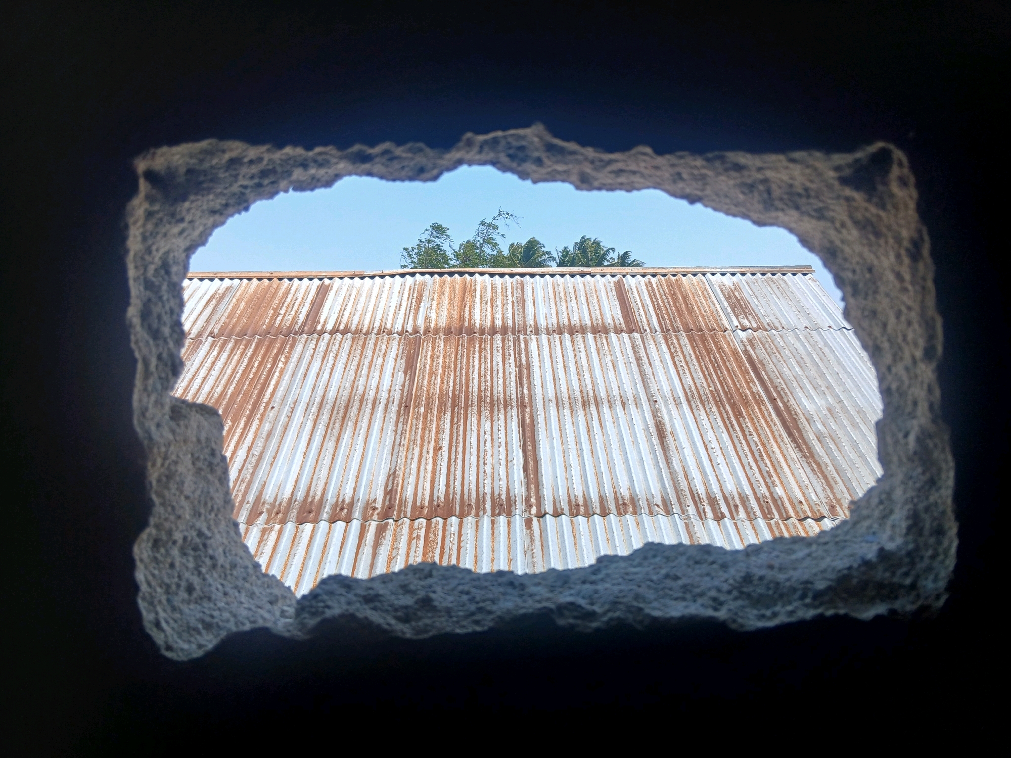 The image shows a view through a jagged hole in a dark wall. The hole is roughly rectangular with irregular, rough edges. Through the opening, a corrugated metal roof with visible rust and streaks of brown is seen, sloping downwards from the top of the frame. Above the roof, a clear blue sky is visible, with the tops of green trees, possibly palm trees, rising above the roofline. The scene appears to be outdoors, possibly in a rural or semi-rural setting, given the nature of the roof and vegetation. The lighting suggests it is daytime, with the sun casting shadows. No people or specific actions are depicted. This scene could be located in various parts of Gorontalo, Indonesia, where such roofing materials and vegetation are common.