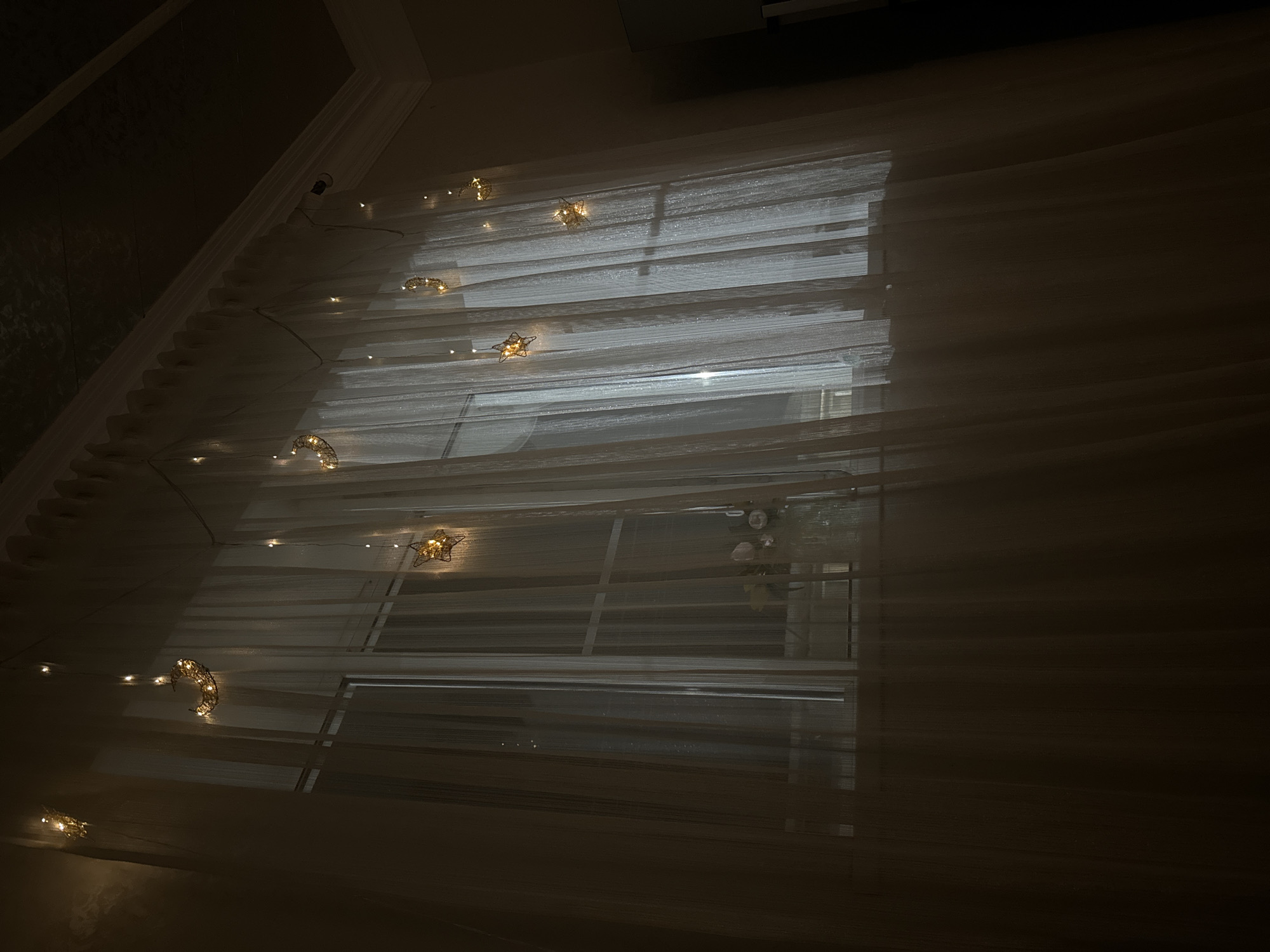 The image shows a window covered by sheer, white curtains. A string of decorative lights with crescent moon and star shapes is draped across the top of the curtains, illuminating them with a warm glow. The lights are a prominent feature against the dark interior and the dimly lit window. A vase with a few flowers is visible on a surface behind the curtains, suggesting the window is in a room. The overall ambiance is calm and dimly lit, with the light from the string decorations creating a gentle illumination. The location is described as Paraul, Russia.