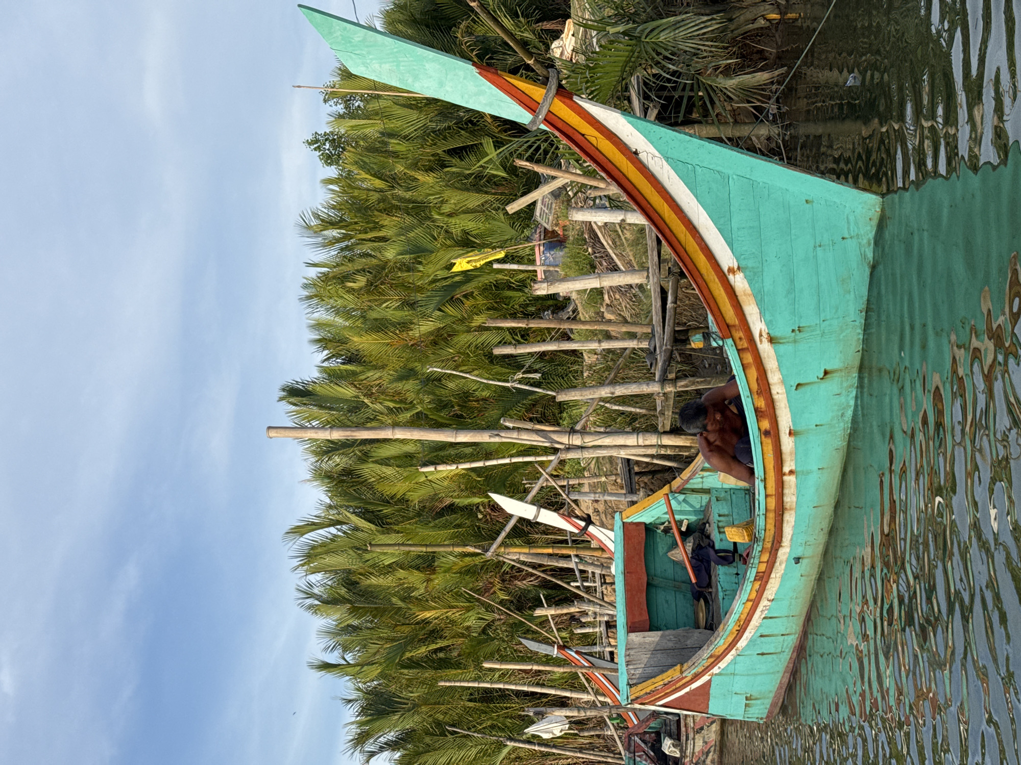 The image depicts a tranquil scene in Reudeuep, Indonesia, focused on a brightly colored fishing boat docked in calm, reflective water. The boat, predominantly teal with orange and yellow accents, has a distinct pointed prow adorned with stripes of orange, yellow, and white. A shirtless man is visible inside the boat, tending to its interior. The background is dominated by a dense wall of lush green palm trees, characteristic of the tropical environment. The sky above is a clear, light blue with a few wispy clouds. Scattered bamboo poles and wooden structures are interspersed among the palm trees, suggesting a riparian or coastal settlement. The water's surface beautifully mirrors the surrounding greenery and the boat itself, adding a serene quality to the composition.