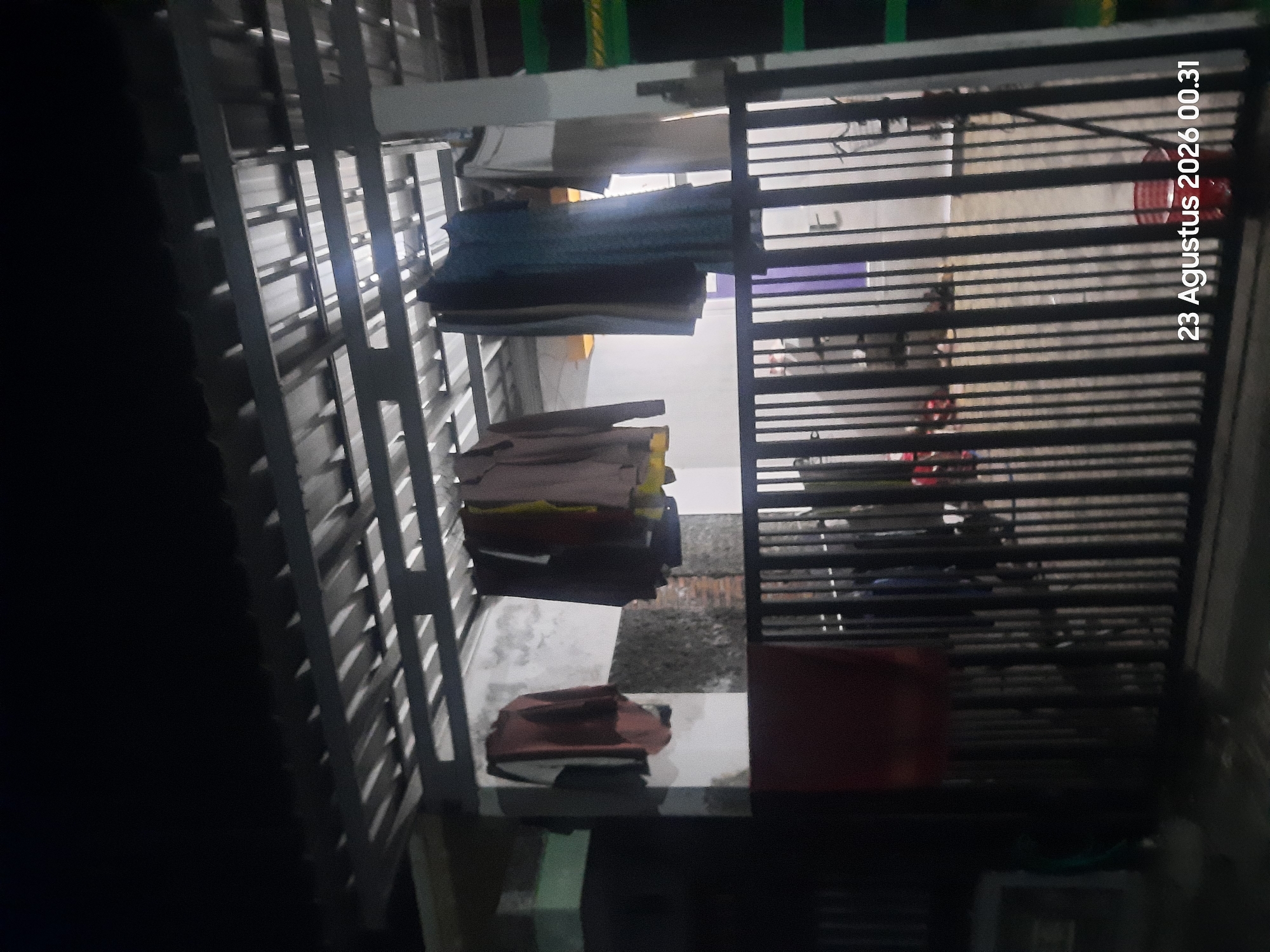 An outdoor scene at night shows clothing hanging on a clothesline under a metal roof structure. The clothing appears to be a variety of garments, some colorful and some neutral. A black metal fence runs across the foreground, partially obscuring the view of the yard behind it. The yard appears to be paved and there are some blurry objects visible in the background, suggesting a residential setting. A red bucket is visible on the ground behind the fence. The date and time stamp "23 Agustus 2026 00.31" is superimposed on the lower right portion of the image. The location is identified as Sukokerto, Indonesia.