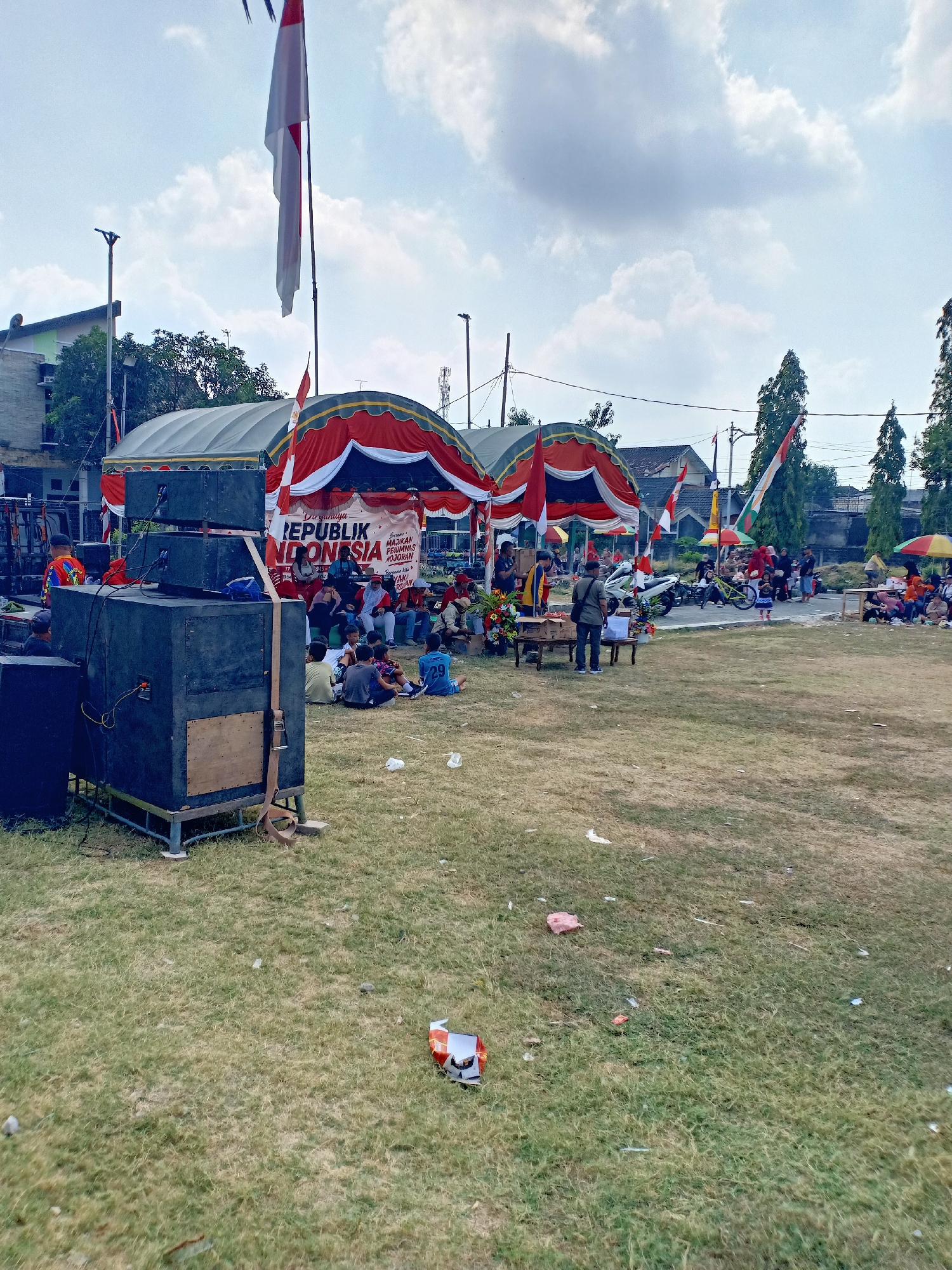 The image depicts an outdoor gathering in Sumber Tlaseh, Indonesia, possibly a community event or celebration. In the foreground, a large black speaker system is set up on a metal stand, positioned on a grassy field. Several Indonesian flags, red and white, are prominently displayed, with one large flag flying from a pole in the upper left of the frame, and smaller flags decorating the tents. Two large tents with red and white awnings are visible in the middle ground, providing shade. A crowd of people, appearing to be a mix of adults and children, are seated or standing near the tents. Some are gathered around a table, while others are seated on the grass. In the background, buildings and trees are visible, along with some parked motorcycles and a few colorful umbrellas. The ground is covered in dry, sparse grass, with some litter scattered about. The sky is overcast with some visible clouds.