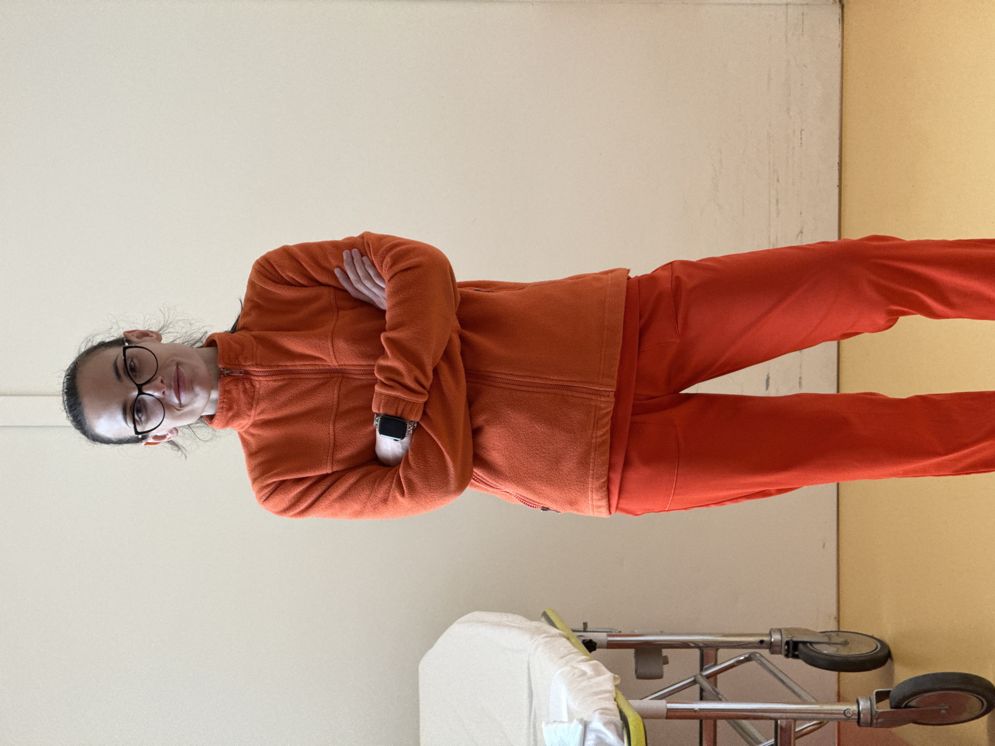 The image depicts a woman in an orange fleece jacket and pants standing in what appears to be a medical facility. She has dark hair pulled back, wears glasses, and has her arms crossed. A gurney with a white sheet and medical supplies is visible near her. The walls are two-toned: a light beige on the left and a pale yellow on the right. The location cannot be determined to be in Pleven, Bulgaria, from the provided information.