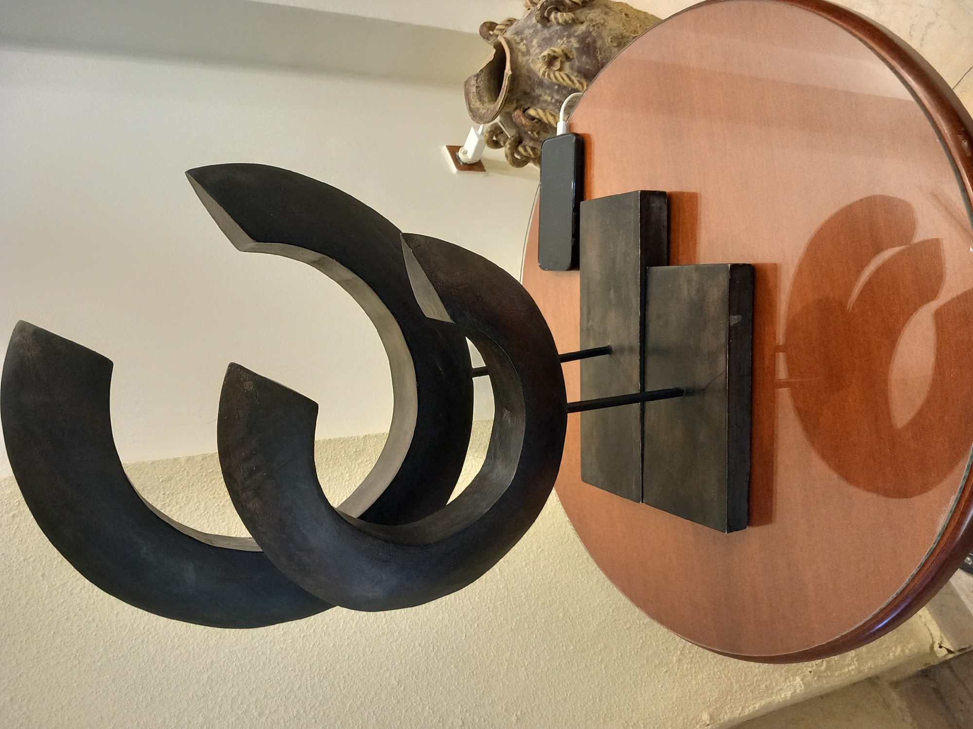 A dark, abstract sculpture is positioned on a round, polished wooden table. The sculpture is composed of two intertwined, thick, curved pieces that appear to be made of a dark, matte material. The base of the sculpture consists of two rectangular black platforms, each resting on thin metal supports.
To the right of the sculpture, a black smartphone is charging, connected by a white cable to a power outlet on the wall. The power outlet is a standard white unit with a brown surround. Behind the smartphone, a large, ornate ceramic vase with rope-like handles and a textured surface is visible. The vase is a muted brown and beige color.
The table is circular with a dark reddish-brown polished surface that reflects the objects placed upon it. The background features a textured light-colored wall and a lighter wall to the right, suggesting an indoor setting. The lighting is neutral, casting soft shadows. The overall scene conveys a sense of modern decor with a touch of antique.