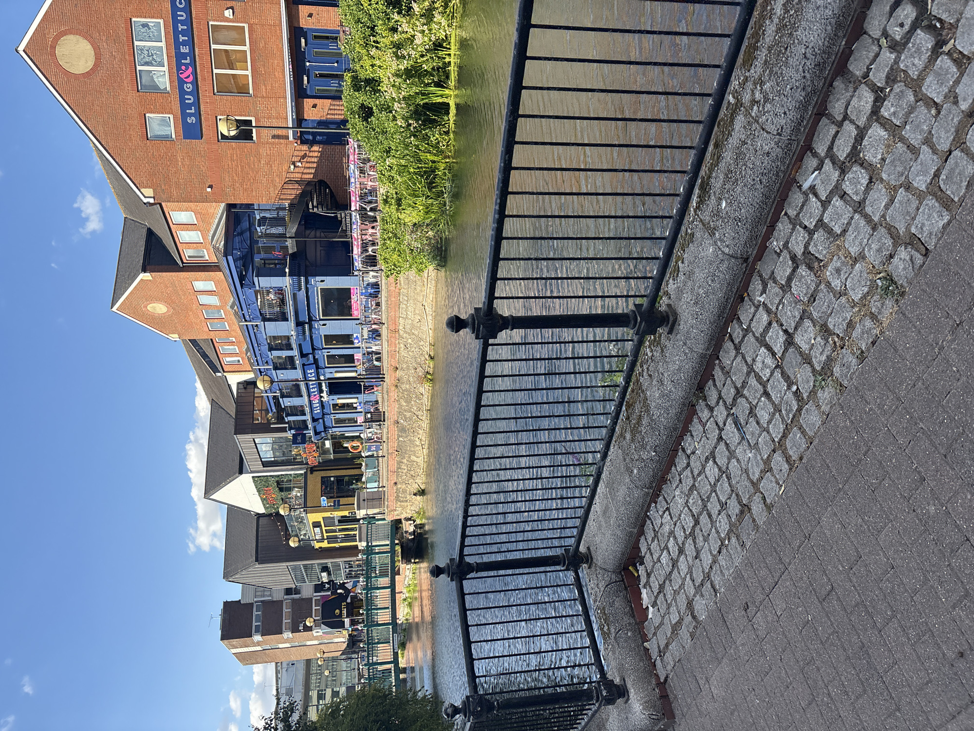 The image depicts a waterside scene in Chelmsford, United Kingdom, likely along the River Chelmer, with a prominent blue building labeled "SLUG & LETTUCE" on the right. The architecture suggests a developed urban area, with brick buildings and modern structures lining the waterfront. The river is calm, reflecting the blue sky and fluffy white clouds. A black metal railing runs along the paved walkway in the foreground, separating it from the river. Beyond the railing, the river flows gently, with lush green foliage growing along its banks. Several other buildings are visible, including a multi-story brick building with multiple windows, and a yellow building with signage, possibly another establishment. A green bridge crosses the river in the distance. There are tables and chairs set up outside the Slug & Lettuce, suggesting an outdoor seating area for patrons. The sun is shining, casting shadows and illuminating the scene with warm light. The overall atmosphere appears peaceful and pleasant, with signs of urban life and leisure.