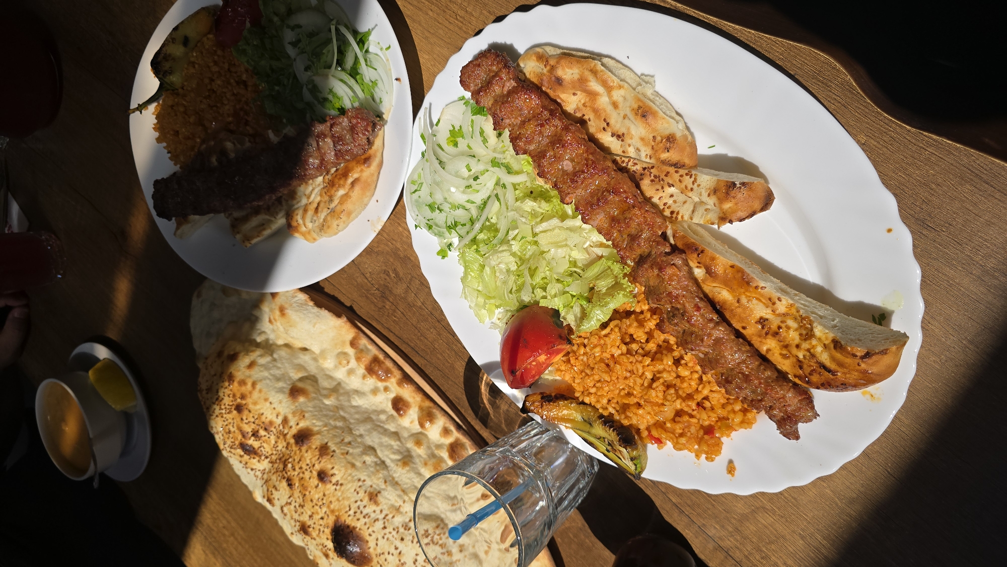 Two plates of Turkish cuisine are presented on a wooden table. The plate in the foreground features two kebabs, bulgur pilaf, a grilled tomato and pepper, shredded lettuce, and sliced onions. The plate in the background also contains kebabs, bulgur pilaf, and a salad. A large piece of flatbread with sesame seeds is situated to the left of the foreground plate. A glass with a blue straw and a small bowl of soup with a lemon wedge are also visible on the table. The lighting suggests this is an indoor setting, possibly a restaurant, with natural light creating strong shadows. The image does not contain any identifiable landmarks or features that would allow for pinpointing the location to Kardzhali, Bulgaria.