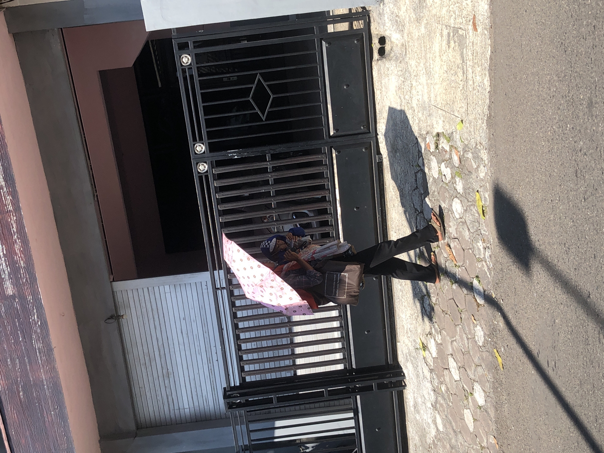 In Trawas, Indonesia, a person walks on a paved sidewalk carrying a child under a pink umbrella. The person is wearing black pants and sandals, and the child is wearing a blue outfit and a patterned head covering. They are walking in front of a black metal gate and a building with a roll-up door. The sun is casting a long shadow on the ground.