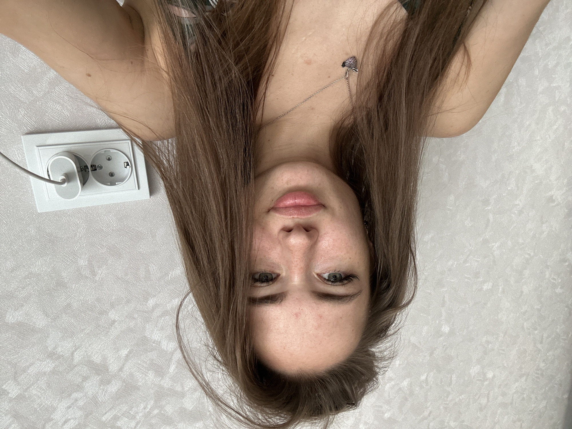 A young woman with long brown hair and light-colored eyes is captured in a close-up selfie against a textured white wall. She is wearing a necklace with a heart-shaped pendant. To her right, a white electrical outlet with two sockets is visible, with a white cable plugged into one of the sockets. The image appears to be taken indoors.