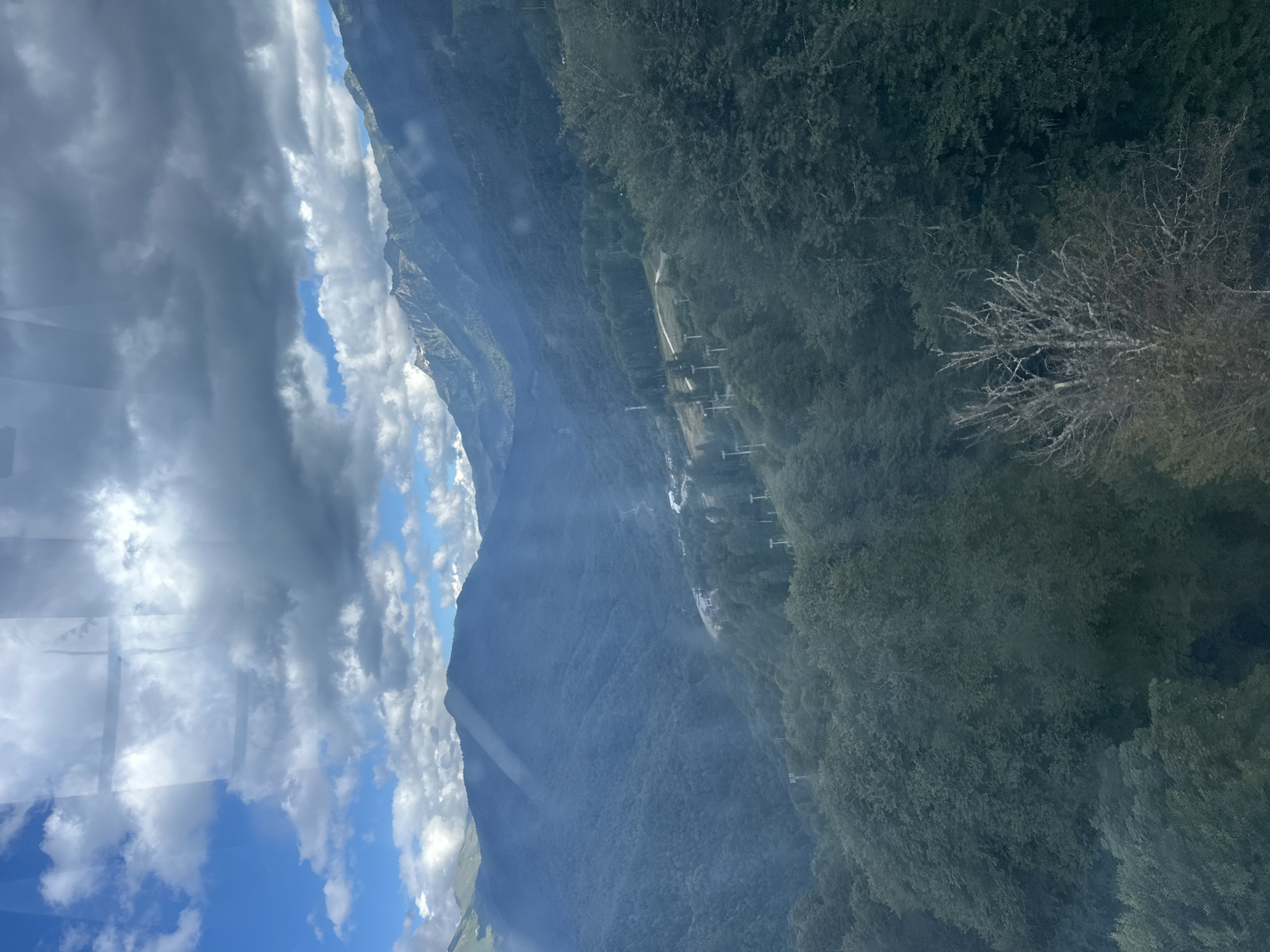 The image captures a panoramic view of a mountainous landscape, likely taken from a cable car, overlooking the ski resort area of Estosadok, Russia. The foreground is dominated by dense, dark green foliage, suggesting a forest or thick vegetation. In the mid-ground, the terrain slopes downwards, revealing a winding road or pathway and a series of ski lift towers, indicative of a ski resort infrastructure. The background features layered mountains that fade into a hazy blue due to atmospheric perspective. The sky above is a dynamic mix of bright blue patches and heavy, dark clouds, with sunlight filtering through in some areas, creating dramatic lighting effects across the mountains. The overall scene conveys a sense of vastness and natural beauty, with the man-made elements of the ski resort integrated into the rugged topography.