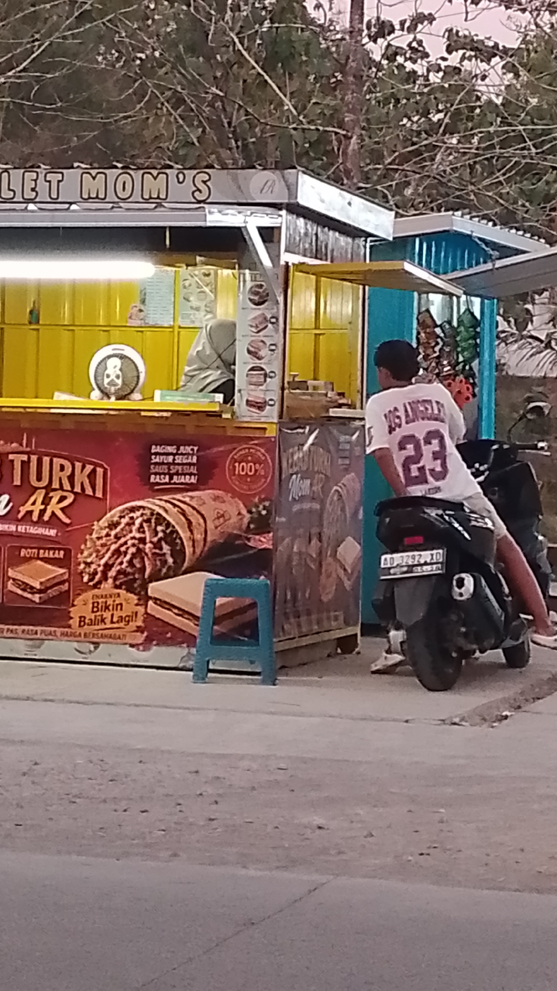 In the city of Karanglangu, Indonesia, a food stall named "LET MOM'S" is visible. The stall has a yellow facade and displays images of food products. A young person wearing a white t-shirt with "LOS ANGELES 23" printed on the back is sitting on a black motorcycle in front of the stall. The motorcycle has a license plate that reads "AD 3292 XD". The stall appears to be selling a product called "TURKI AR" which is described as having "RASA JUARA!" (champion taste). There are also images of roti bakar (toast) on the stall's advertisement. The environment appears to be outdoors, with a dirt or gravel road in the foreground and trees in the background.