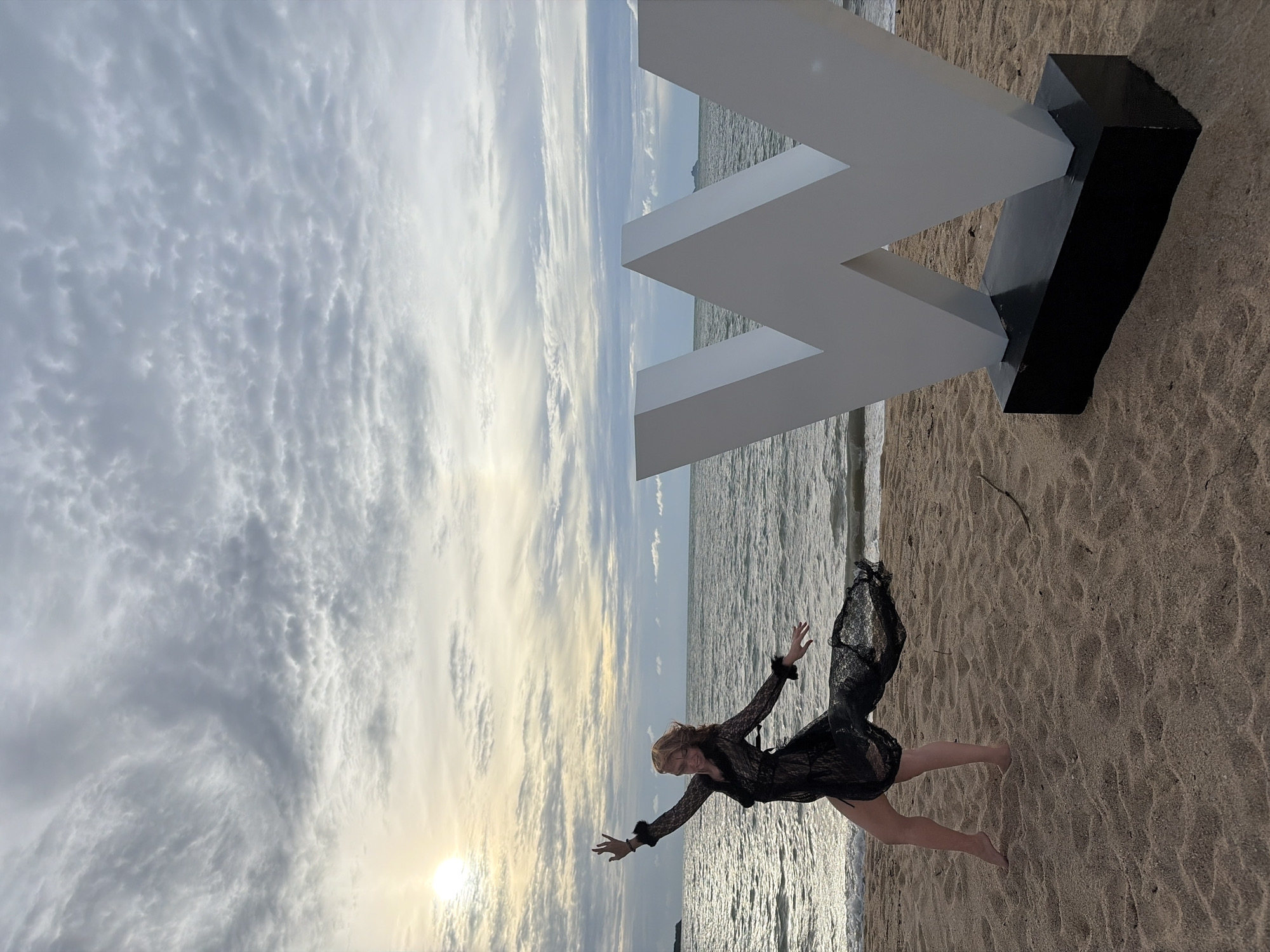 A woman in a black lace dress dances on a sandy beach in Ko Samui District, Thailand. The sun is setting, casting a warm glow on the scene. A large, white "W" sculpture stands to the right of the woman. The ocean stretches out behind them, with a cloudy sky overhead.