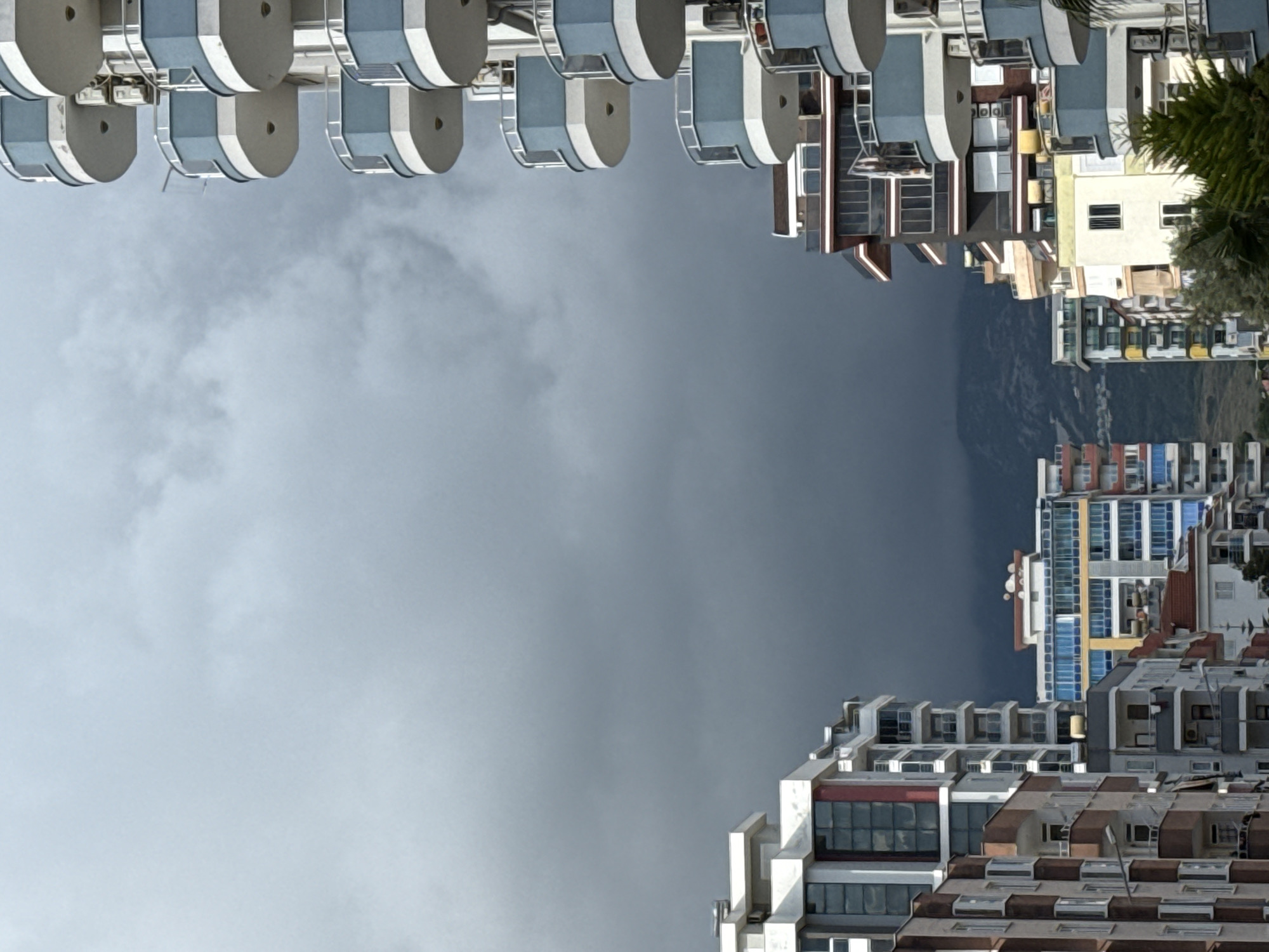 Here is a detailed description of the image:
The image depicts a city scene with several buildings and a cloudy sky. The perspective is somewhat unusual, as the image appears to be inverted or viewed from a low angle, making the buildings seem to tower above. The buildings have a mix of architectural styles, some with rounded balconies, others with rectangular windows, and varying roof types.
The sky is overcast with heavy clouds, creating a moody atmosphere. The buildings are located in a city. Some mountainous terrain is visible in the background.