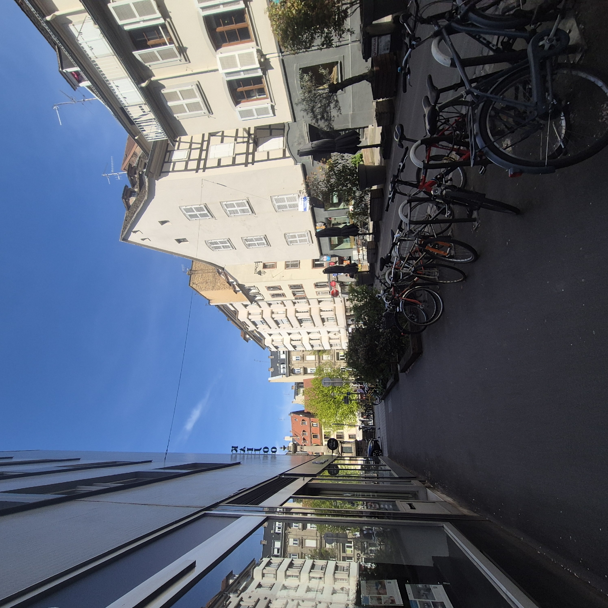 This is an eye-level, outdoor shot of a narrow street in Strasbourg, France, with buildings on both sides and bicycles parked along the right. The sky is a clear, deep blue. The buildings are multi-story structures with various architectural styles, including visible timber framing and white window shutters. Some buildings are reflected in the windows of the lower building.
On the right side of the street are several parked bicycles.
In the distance, the street appears to lead to an open area with more greenery and buildings. A sign with the letters "KYOTO" is visible in the distance. The overall scene appears to be taken on a sunny day, with bright light illuminating the buildings and street. The street appears to be a relatively quiet and well-maintained area of the city.