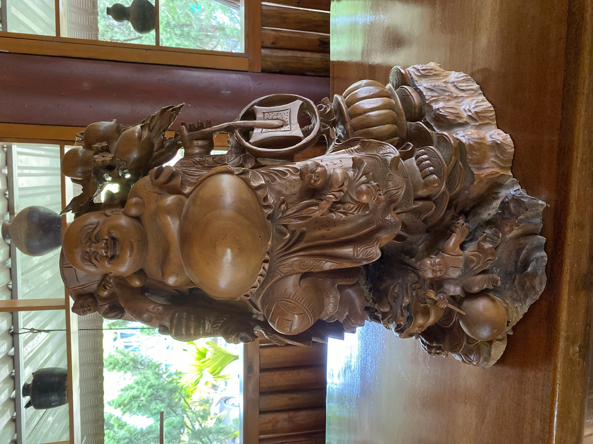 The image shows a large, intricately carved wooden statue of a jovial, rotund figure, likely representing the Budai or "Laughing Buddha". The statue is predominantly made of polished, light brown wood and is positioned on a wooden table. The Budai figure is depicted with a wide smile, a prominent belly, and is adorned with flowing robes. Around him are various carved objects including gourds, fruits, and smaller figures of children. One hand holds a Ruyi scepter, a ceremonial object, while another appears to be resting on a large sack. The carving is detailed, with textures and patterns visible on the robes and surrounding elements.
The setting appears to be indoors, with hints of natural light coming from windows. Through the windows, lush green foliage and trees are visible, suggesting an outdoor environment with abundant greenery. The interior elements include wooden paneling and some decorative ceramic gourds hanging from the ceiling, visible through the window. The lighting suggests it is daytime, with soft, natural light illuminating the statue. The overall impression is one of peace, prosperity, and good fortune, often associated with the Budai figure. The scene is static, with the statue being the sole subject of focus. No visible text is present in the image.